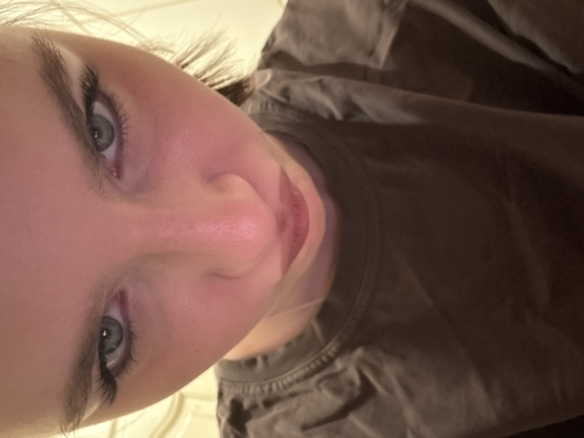 A close-up, tilted, and somewhat obscured image presents a portion of a person's face and upper torso. The face is fair-skinned with grey eyes and visible eyelashes enhanced with mascara. The skin appears clear, with a slight pinkish tone. The person's nose and mouth are centered in the frame, with the mouth slightly open. The person is wearing a dark-colored t-shirt. The background is out of focus, revealing a partial view of what looks like a white surface. There is no discernible indication of the location being Vsevolozhsk, Russia.