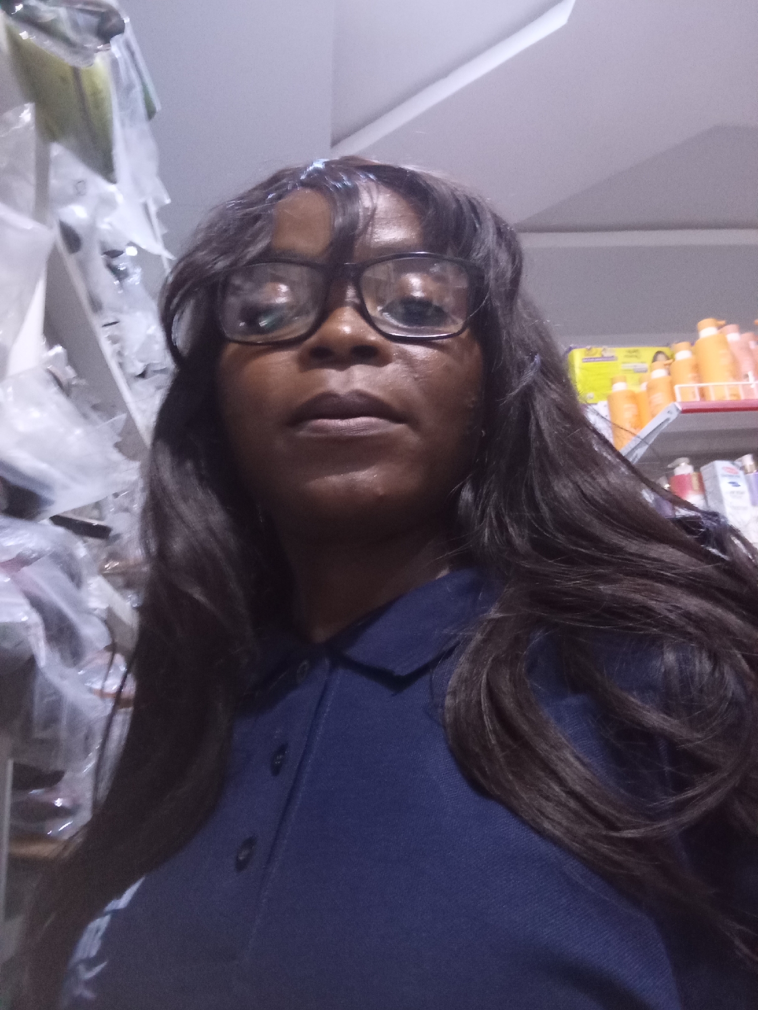 A medium shot, close-up selfie depicts a Black woman with dark hair and glasses. She is wearing a dark blue collared shirt. The background appears to be a retail environment with shelves holding various products, including orange bottles and clear plastic-wrapped items. The lighting is dim and appears to be artificial. The specific location in Howayi, Nigeria cannot be determined from the image.