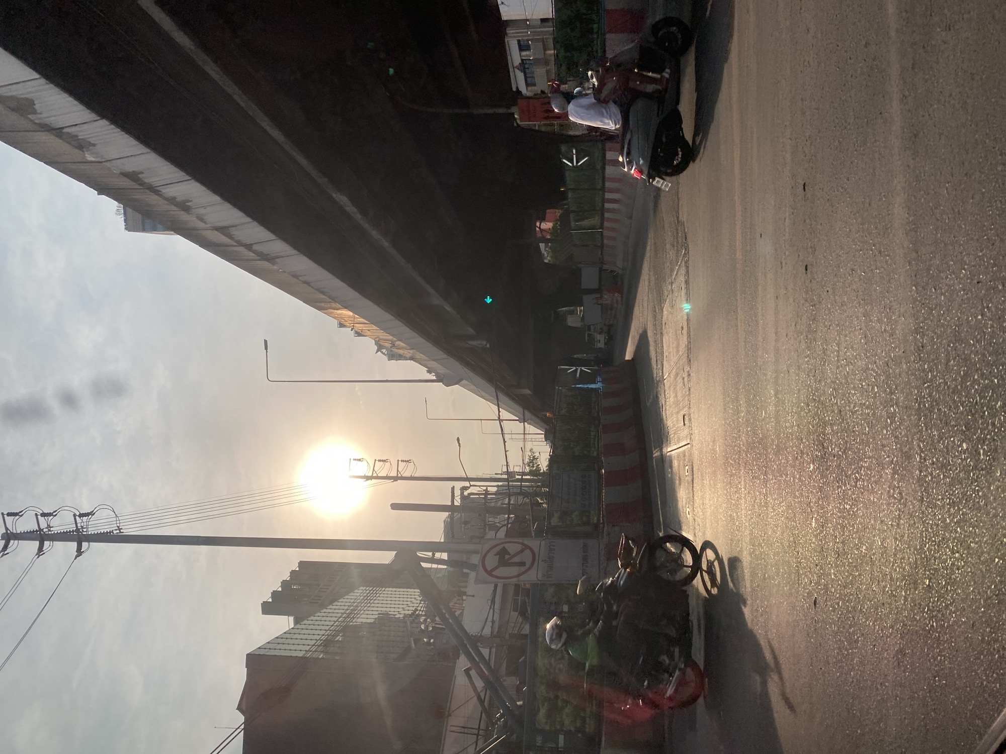 The image depicts a street scene in Bangkok, Thailand, taken from beneath an elevated structure. The sun shines brightly in the distance, partially obscured by buildings and utility poles. On the right side of the frame, two motorcycles are parked on the side of the road, one with a rider. A red and white striped barrier runs along the edge of the road. Traffic signs are visible amidst the urban clutter. The elevated structure above casts a shadow over the street, creating a contrast between light and darkness. The overall environment is urban, with buildings and infrastructure elements dominating the view.