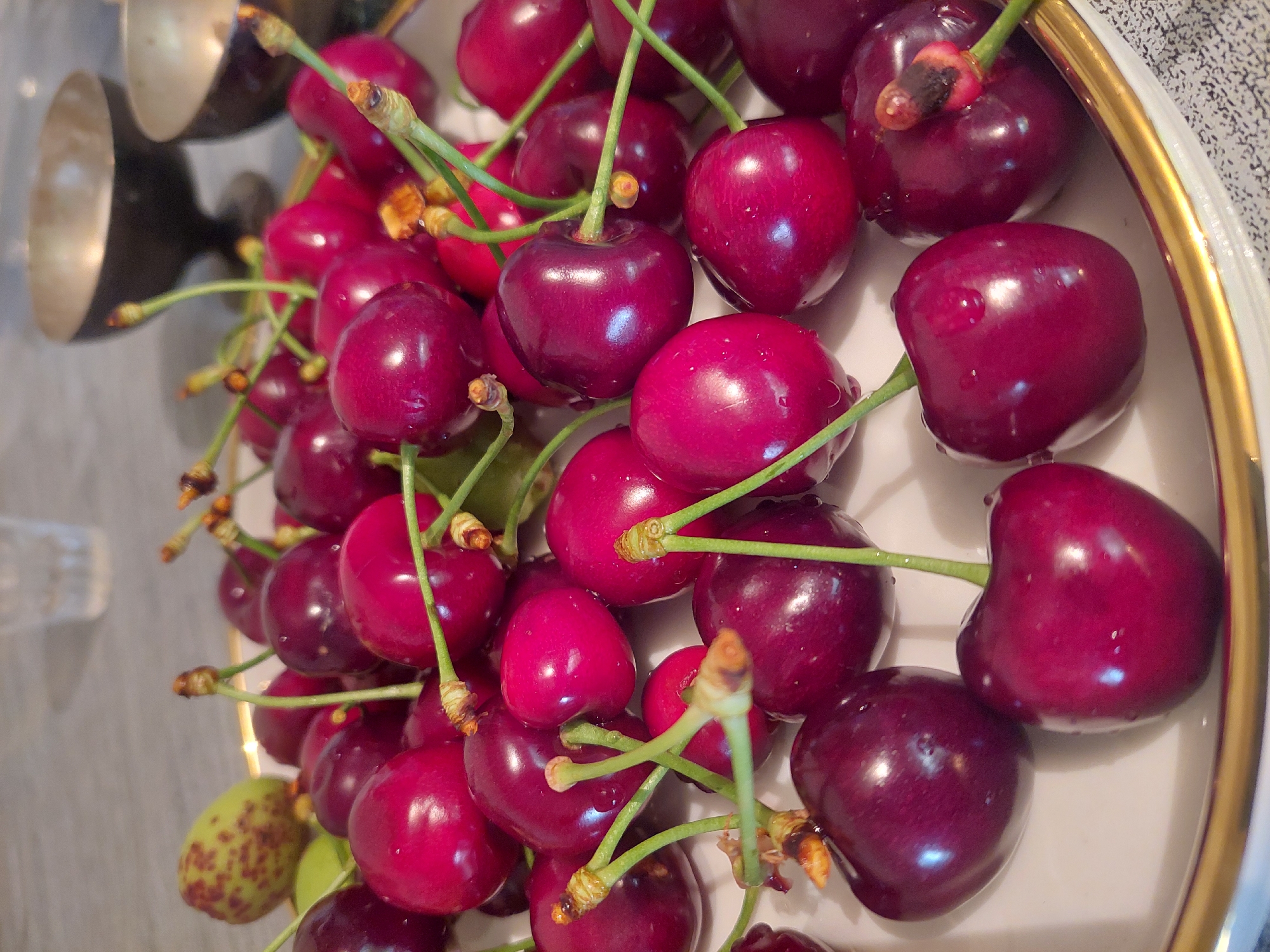 The image presents a close-up, slightly tilted overhead view of a generous pile of dark red cherries resting on a white plate with a prominent golden rim. The cherries are a deep, glossy burgundy color, many displaying small water droplets on their skin, indicating freshness or recent washing. Each cherry is connected to a vibrant green stem, with some stems still attached to multiple fruits, forming small clusters.
In the upper left portion of the image, reflections from what appear to be metallic bowls or utensils are visible, suggesting an indoor setting. Towards the bottom left, partially obscured, is a single, light green fruit with reddish-brown speckles, distinct from the cherries.
The composition focuses tightly on the fruit, emphasizing their rich color and dewy appearance. No human activity or specific time of day is discernible. The image lacks any visible text. While the location context of Areni, Armenia, is provided, there are no specific visual cues within the image itself to confirm this detail.