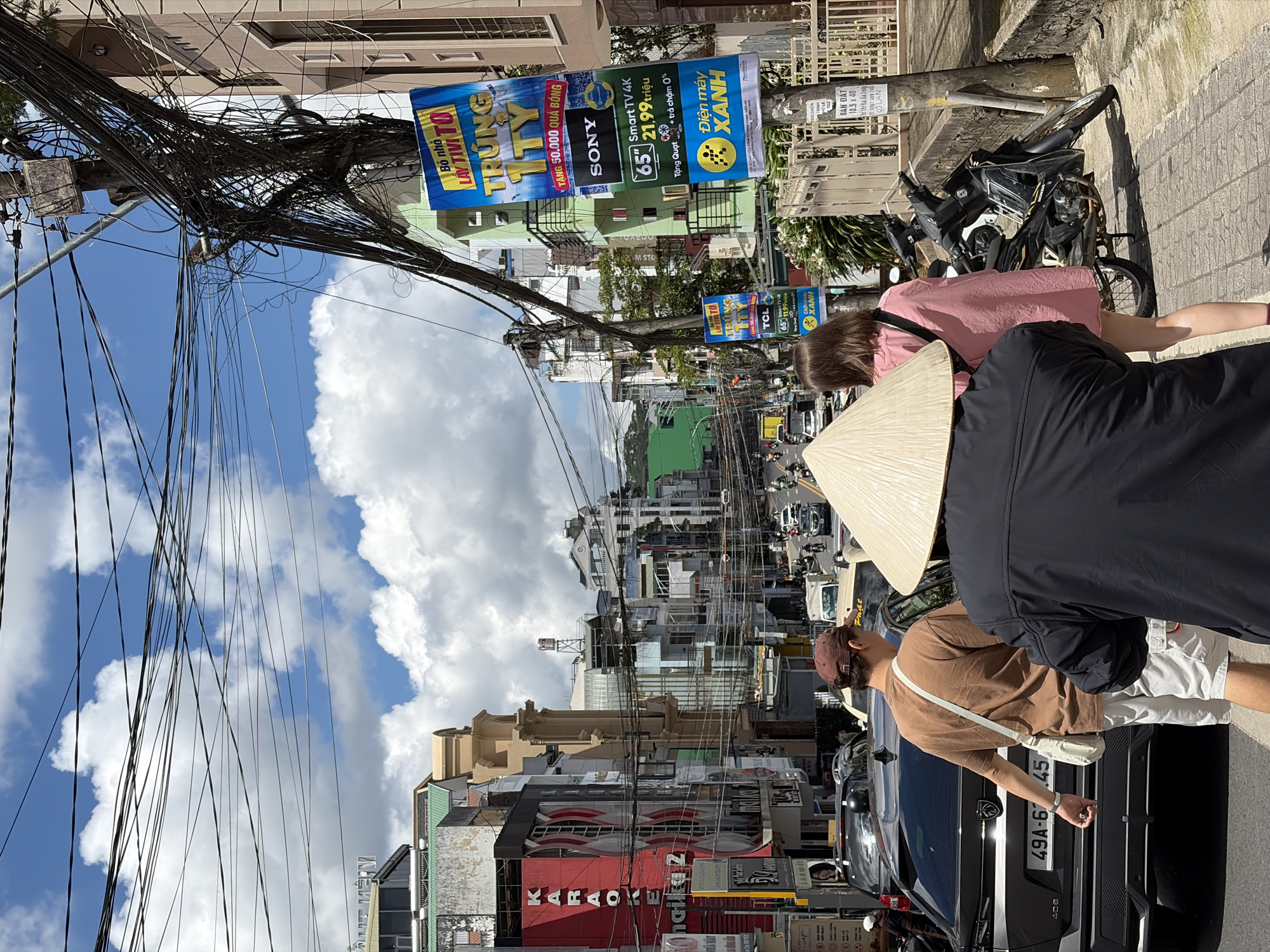 The image captures a street scene in Cam Ly, Đà Lạt, Vietnam, characterized by a dense network of overhead electrical wires and a vibrant urban environment. In the foreground, three individuals are visible. The person furthest back and to the right is wearing a conical hat and a black jacket. To their left, a woman in a pink dress walks away from the camera. In the center, a man wearing a brown t-shirt and white shorts, with a brown baseball cap, walks away from the camera, carrying a white bag slung across his body. Behind him, a dark grey car with license plate "49A-621.45" is parked.
The street is lined with multi-story buildings of varying architectural styles and colors, with prominent signage including "KARAOKE" and advertisements for Sony and TCL products, indicating commercial activity. The buildings are adorned with balconies and windows, and electrical conduits are visibly tangled around utility poles. Further down the street, traffic, including cars and numerous motorbikes, is present. The sky is bright blue with scattered white clouds. A metal fence and a concrete wall are visible on the right side of the image, bordering a sidewalk. A black motorbike is parked on the sidewalk. The overall impression is of a busy, sunlit street in an Asian city, with a common element of heavily organized electrical wiring.