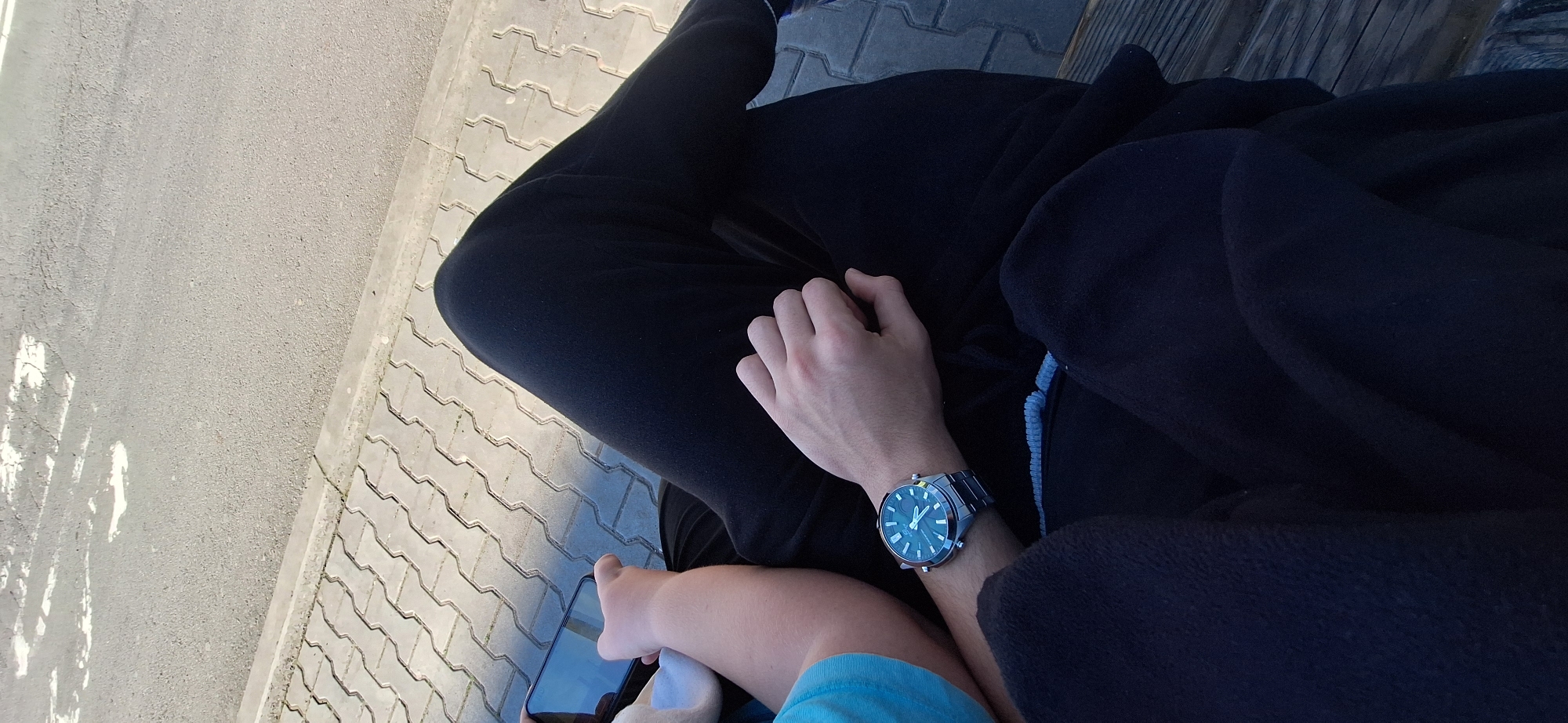 A high-angle shot depicts a person seated, wearing black pants and a dark blue hoodie, with a blue t-shirt visible underneath. The person has a silver watch on their left wrist and is holding a smartphone in their left hand. The setting appears to be outdoors on a paved surface made of interlocking concrete bricks. A section of what looks like an asphalt road or walkway is visible on the left. The location may be in the city of Stara Zagora, Bulgaria, but there is no specific feature to verify that from the image.