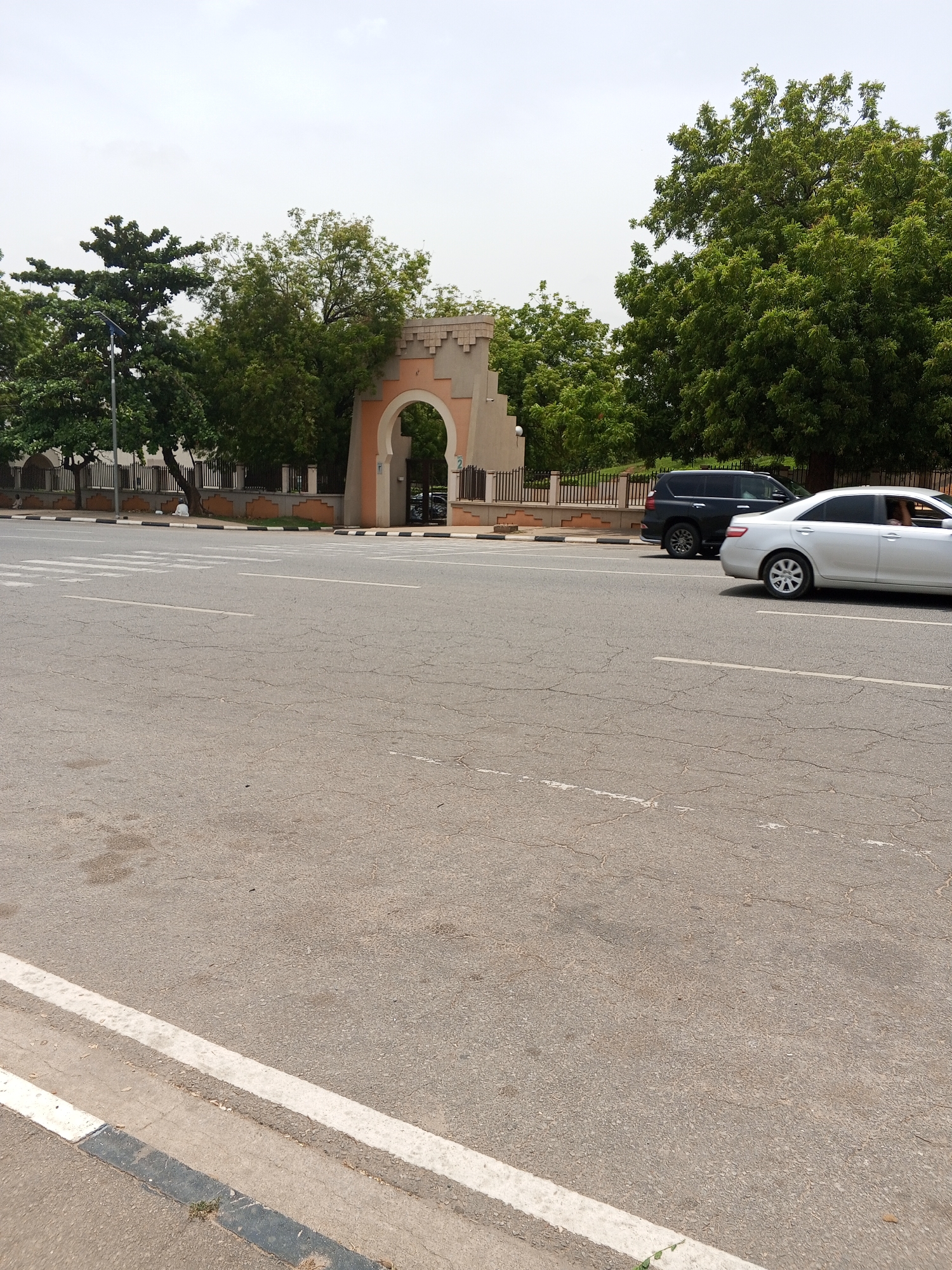 The image shows a street scene in Abuja, Nigeria, featuring a distinctive archway-shaped entrance with a pink and beige facade. The archway is flanked by trees and a low fence. Two vehicles, a silver sedan and a black SUV, are visible on the road. The road itself is cracked and shows signs of wear, with a white lane marker along the curb. The sky is overcast, suggesting a dull or cloudy day. The composition is centered on the road and the archway, providing a clear view of the architecture and traffic.