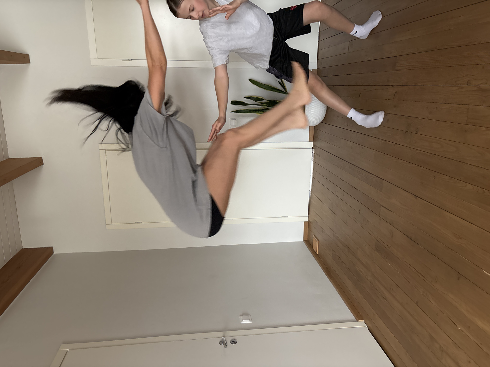 Two people are in the interior space of a building. One person with long dark hair, wearing a gray shirt and black shorts, is mid-air, performing a flip. The other person, with shorter hair and wearing a white shirt with black shorts and white socks, is standing near a potted plant. The room has wooden floors and white walls. The photo is oriented sideways. The location is not discernible.