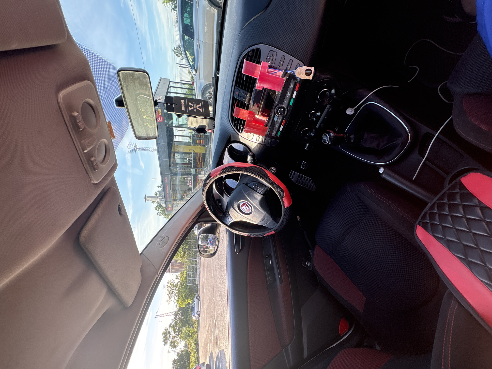 The image captures the interior of a Fiat car, viewed from the driver's seat. The car is parked in a parking lot with a casual dining establishment visible in the background, suggesting a roadside location. The dining establishment appears to be a grill bar with outdoor seating and a drive-through service. The sky is clear and blue, indicating good weather. The interior of the car is decorated with red and black accents, including the steering wheel cover and seat upholstery. The dashboard features a car stereo, air conditioning vents, and a gear shift. A phone mount with a red attachment is visible on the dashboard. The overall scene conveys a sense of everyday life, possibly during a stop at a rest area or a quick meal. The location is identified as being in the city of Vratsa, Bulgaria.