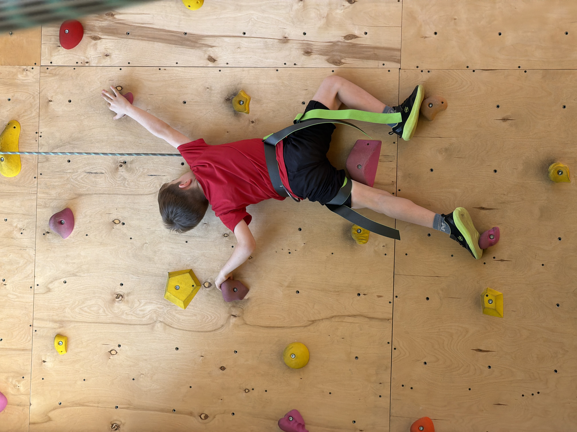 A boy in a red shirt and black shorts is climbing an indoor climbing wall. He is wearing climbing shoes and a safety harness with green straps. The climbing wall is made of light-colored wood panels with various colored climbing holds attached. The boy is extending his arms and legs to reach the holds. A rope is visible along the left side of the wall. The location is an indoor climbing facility, likely in a city but not necessarily Izhevsk, Russia, as that information is not visually discernible.