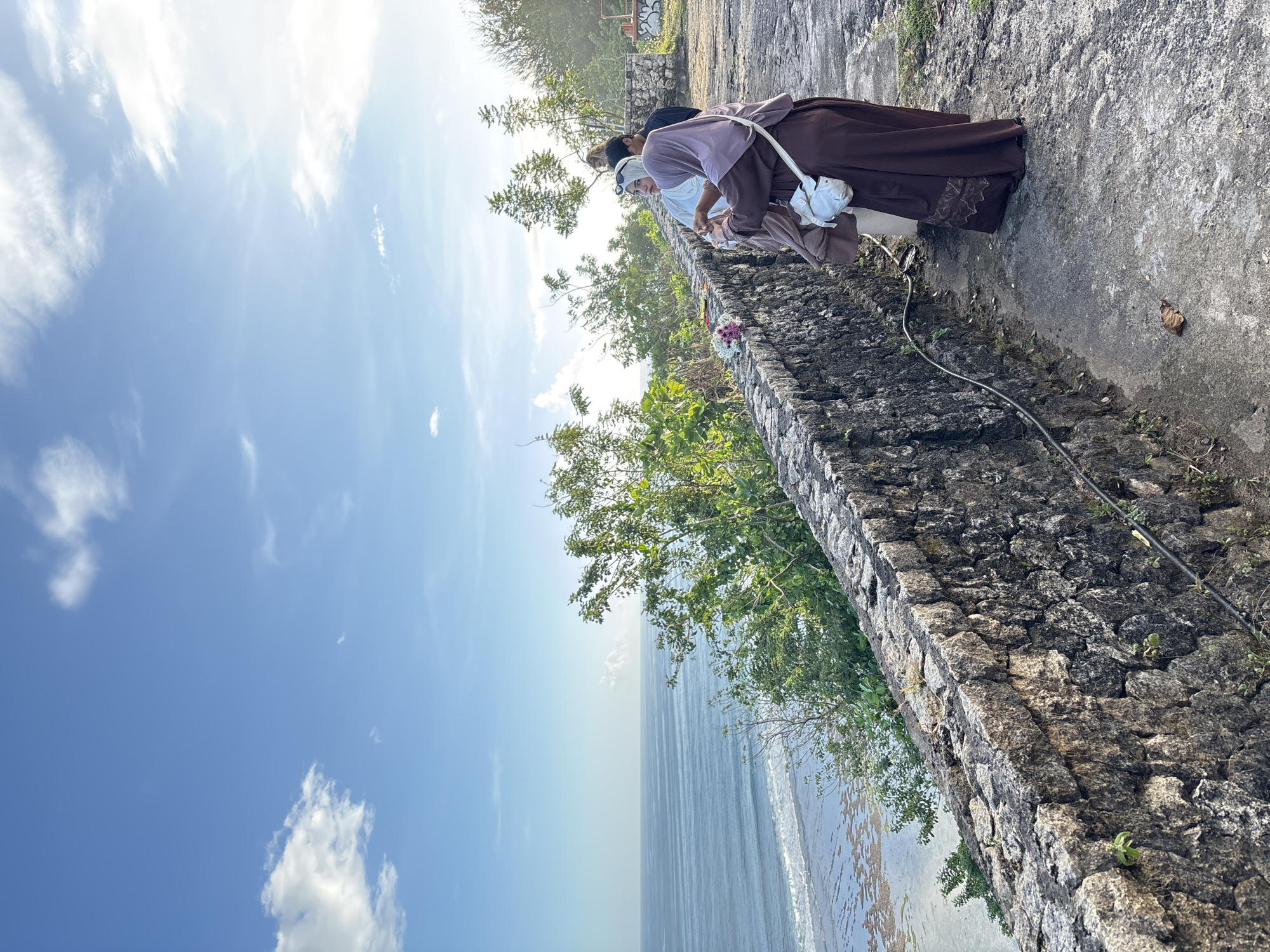 Two people stand beside a stone wall, peering towards the ocean in Kuta Selatan, Indonesia. The person closer to the viewer wears a long brown and mauve garment, with a white bag over their shoulder and a white head covering. The person behind them is wearing a dark top and appears to be looking over the wall as well. The wall is made of rough, dark stone. Lush trees and plants grow on the other side of the wall and in the background. Beyond the wall is the ocean. The sky is a clear blue with a few scattered clouds.