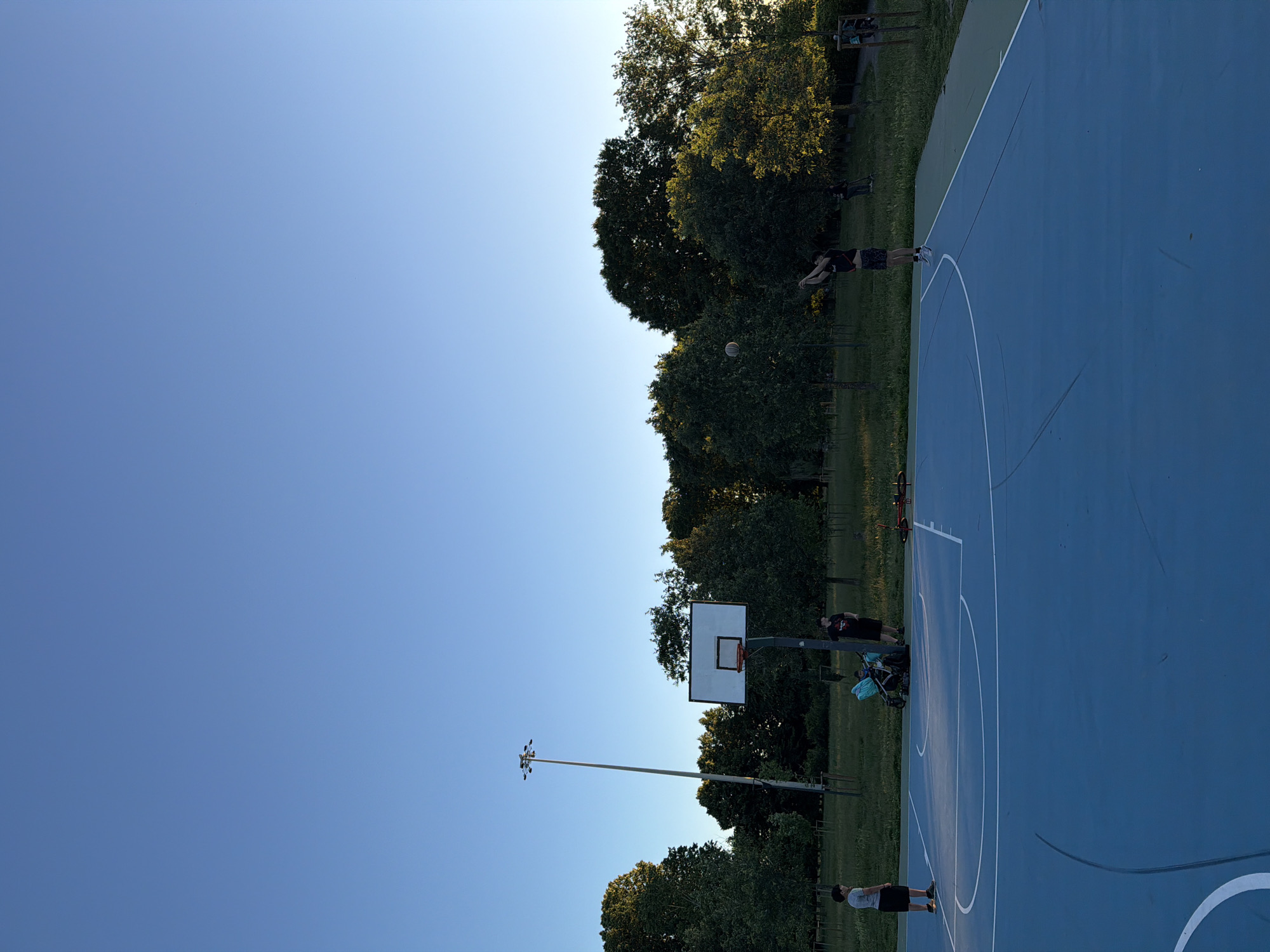 The image shows an outdoor basketball court with a blue surface, located in what appears to be a park setting. There are trees lining the edge of the court and surrounding areas. Several people are present, some playing basketball and others standing or watching. There is a basketball pole visible, along with a light pole extending into the sky. The sky is blue and the lighting suggests it is daytime. Although the location cannot be confirmed, the basketball court appears to be located in Milan, Italy.
