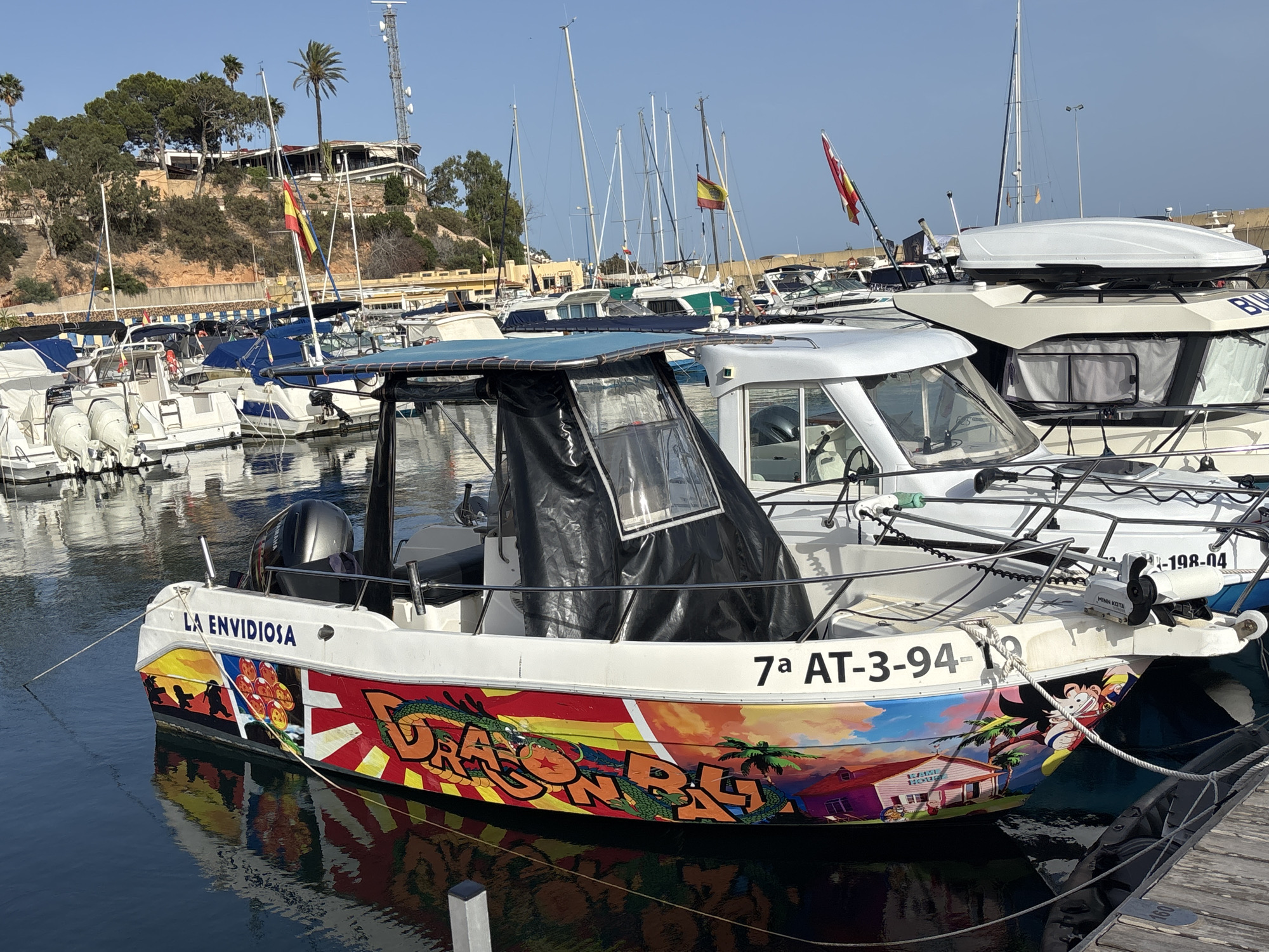 The image shows a fleet of boats docked in a marina in Urbanización Cabo Roig, Spain. The primary focus is a small white motorboat with a black canopy, adorned with colorful Dragon Ball Z-themed artwork on its hull, including characters like Goku and Shenron, along with palm trees and a house. The boat is named "LA ENVIDIOSA" and displays the registration number "7a AT-3-94". Other boats, including larger white vessels and sailboats, are visible in the background, with Spanish flags flying from some of them. A hill with buildings and communication towers overlooks the marina. The water is calm and reflects the boats and the sky.