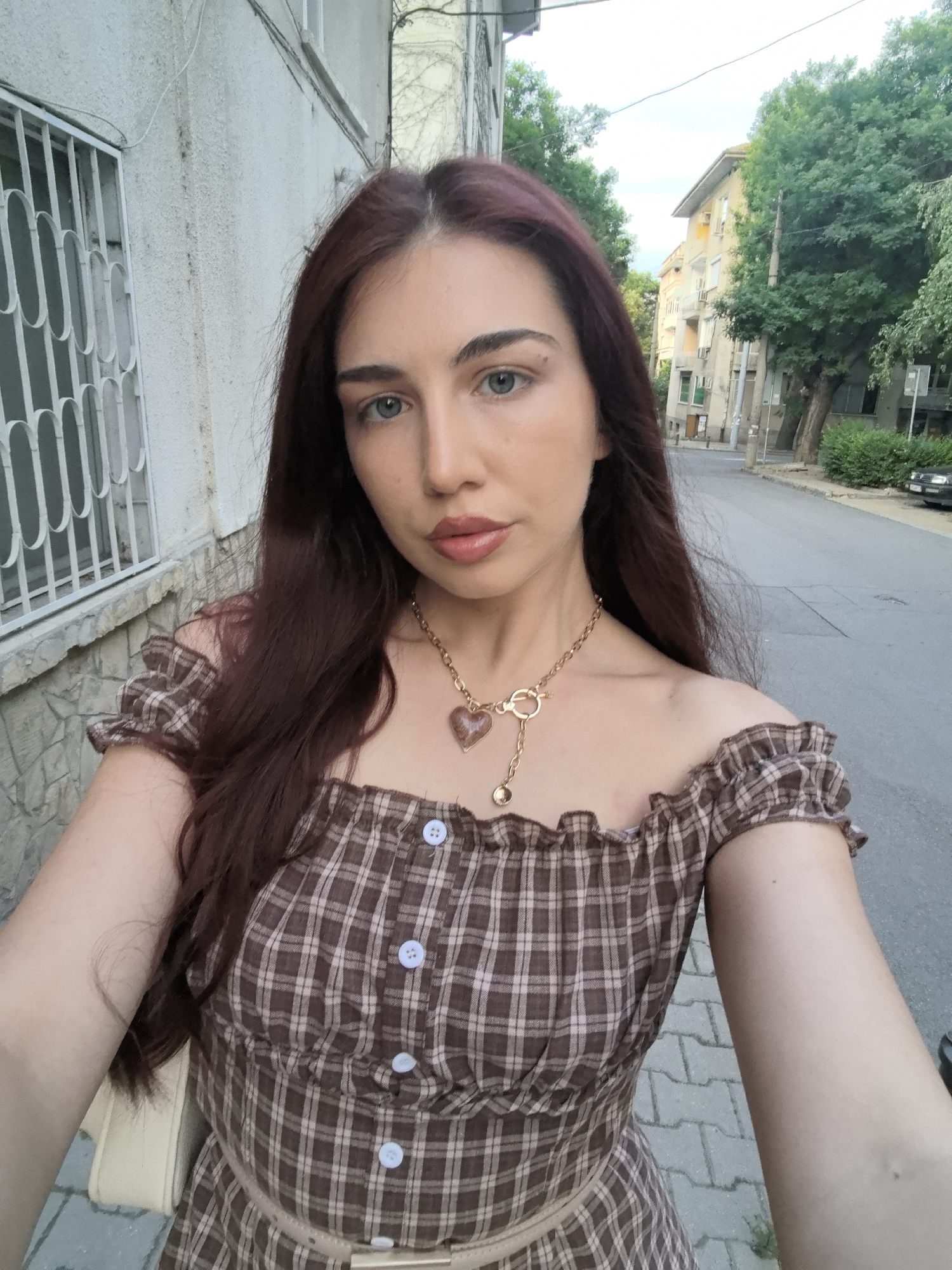 A young woman with long, dark red hair and light blue eyes stands on a paved sidewalk. She is wearing an off-the-shoulder brown and white plaid dress with ruffled sleeves and buttons down the front. A gold necklace with a heart-shaped pendant hangs around her neck. She is holding a light-colored bag in her left arm. In the background, there is a building with a white-barred window, trees, and a street with parked cars. The location is the city of Stara Zagora, Bulgaria.