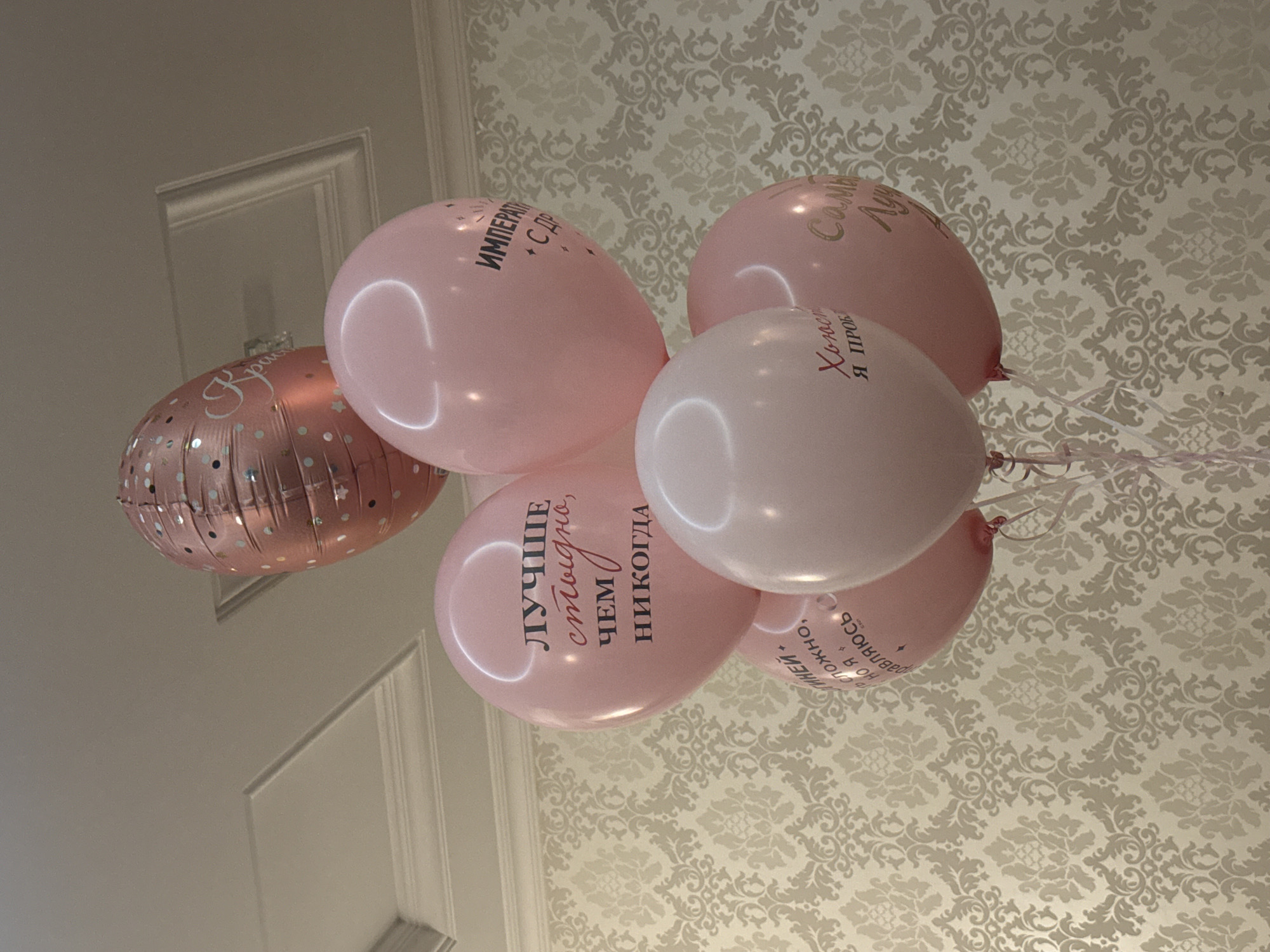 The image shows a bouquet of pink and rose gold balloons indoors, likely a room with patterned wallpaper and a white ceiling. One balloon is rose gold with a polka dot pattern and text that appears to say "Prinsessa". Several pink balloons have text written on them in Russian. One balloon says "ЛУЧШЕ стыдно, чем никогда" (Better to be ashamed than never). Another balloon has text that is partially obscured but seems to include "ИМПЕРАТОР" and "С ДР". A third pink balloon has text that says "Хочу я проб". A fourth pink balloon has text that says "Самыи Лучши". The balloons are attached to ribbons and are floating upwards. There is no indication of the city of Novosibirsk, Russia, or any specific location.