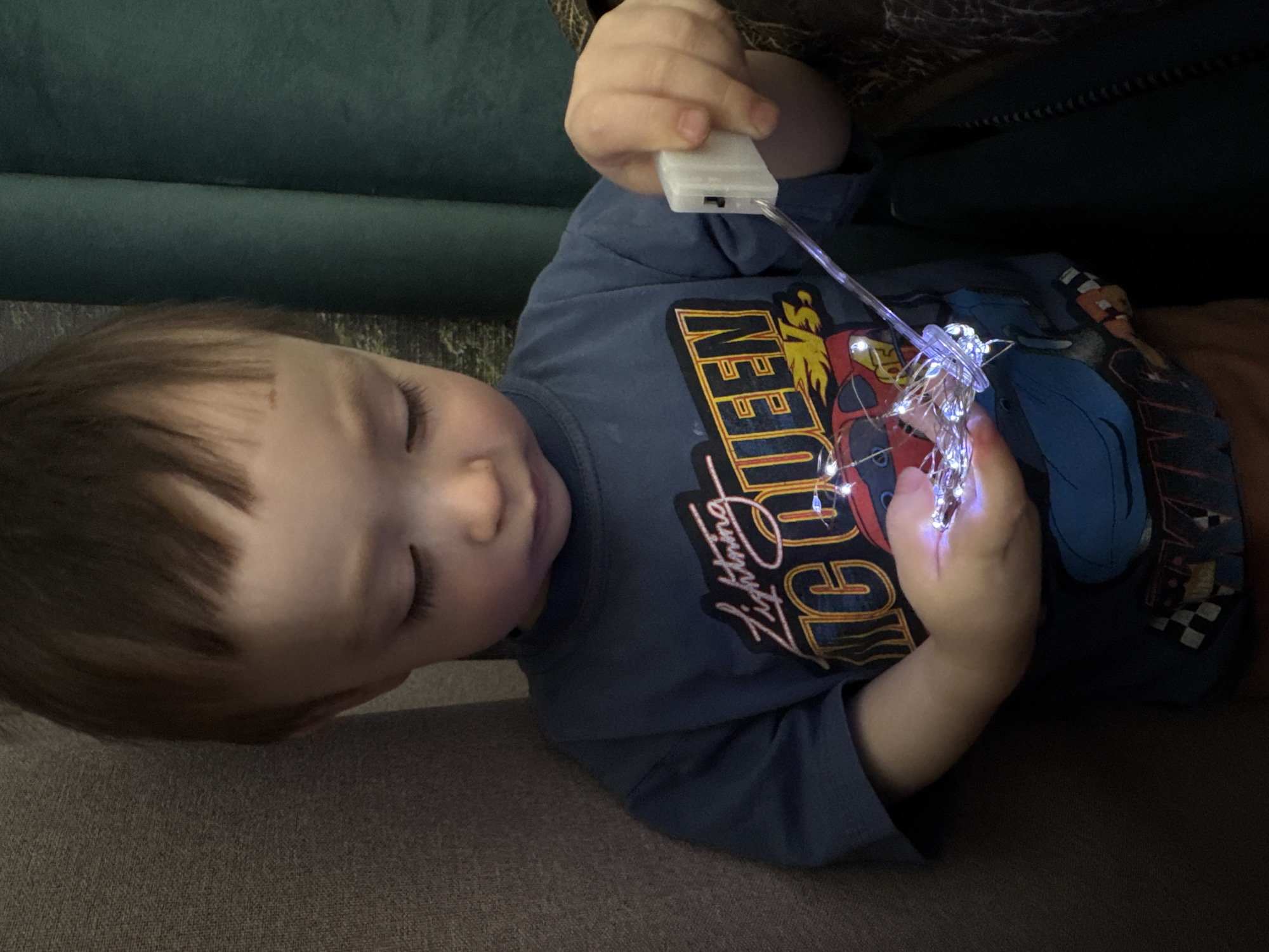 A young child, likely under five years of age, is lying down. The child is fair-skinned with brown hair and appears to be resting with closed eyes. The child is wearing a blue long-sleeved shirt with a "Lightning McQueen" graphic on the front. The child is holding a string of small, illuminated fairy lights in their hands, with the battery pack visible in the child's left hand. The background consists of a dark fabric or upholstery, possibly a couch or cushion. There is no indication of the location of Izhevsk, Russia in the image.