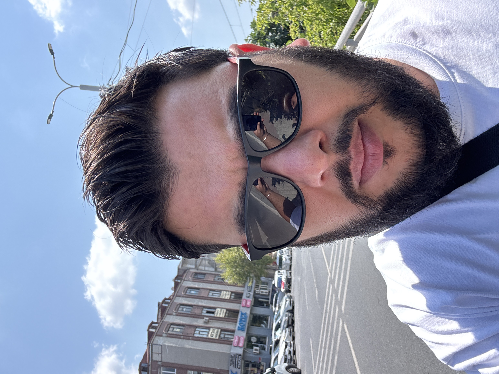 A man is taking a selfie outdoors in Gungoren, Turkey. He is wearing a white t-shirt and black sunglasses. The background shows a street with cars, buildings, trees, a streetlamp, and a blue sky with clouds. His sunglasses reflect a person holding a cell phone, presumably taking the picture, and the surrounding environment. The man has dark hair and a beard.