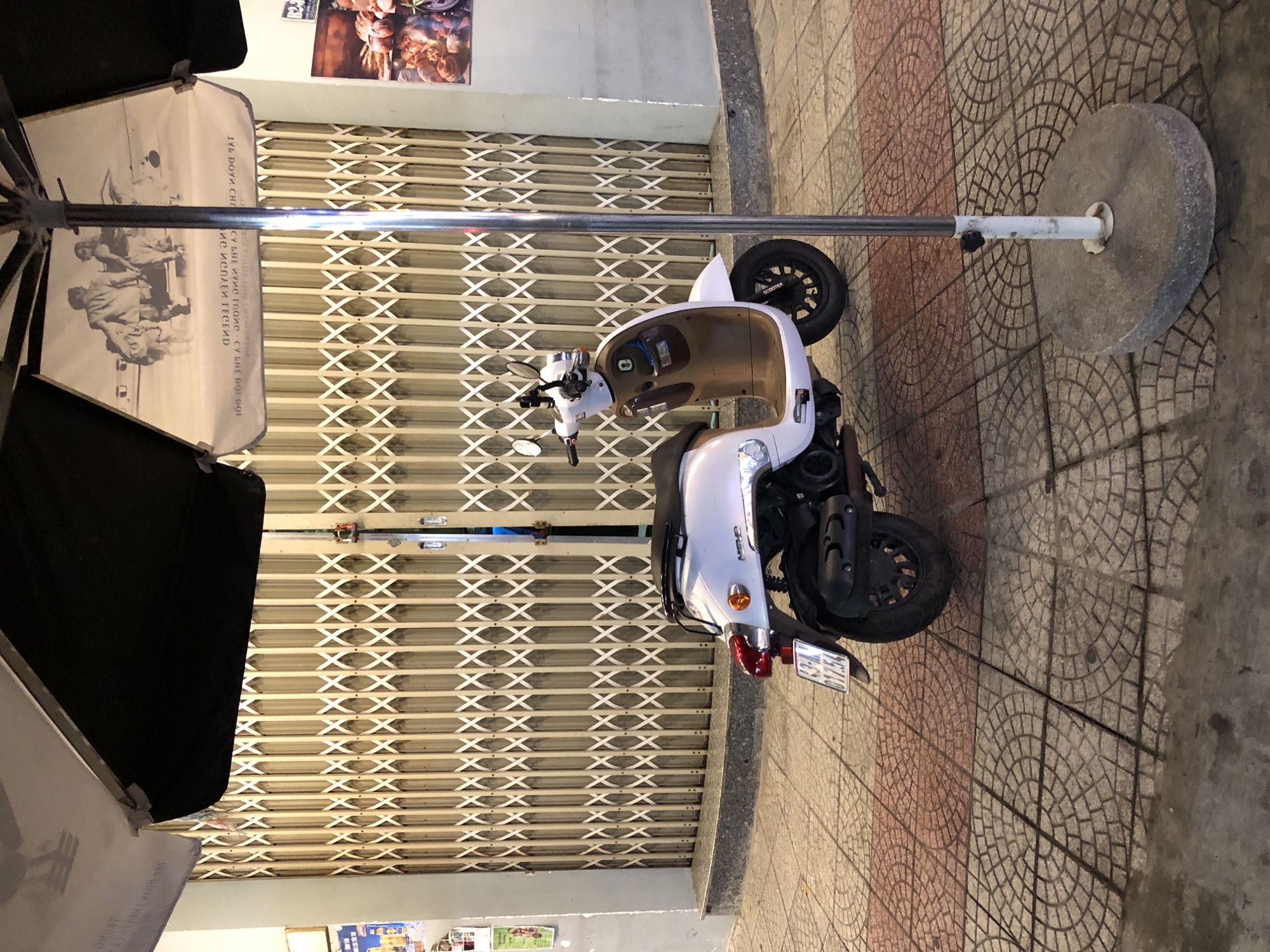 A white scooter with a brown interior is parked on a sidewalk in front of a metal roll-up gate. The scooter has a license plate that reads "63-A 017.54". An umbrella with a black top and a printed image on its underside is angled over the scooter, held up by a metal pole with a concrete base. The sidewalk is paved with patterned tiles. A poster is visible on the wall to the left of the gate. The scene appears to be in a city, possibly in Đà Nẵng, Vietnam, given the context.