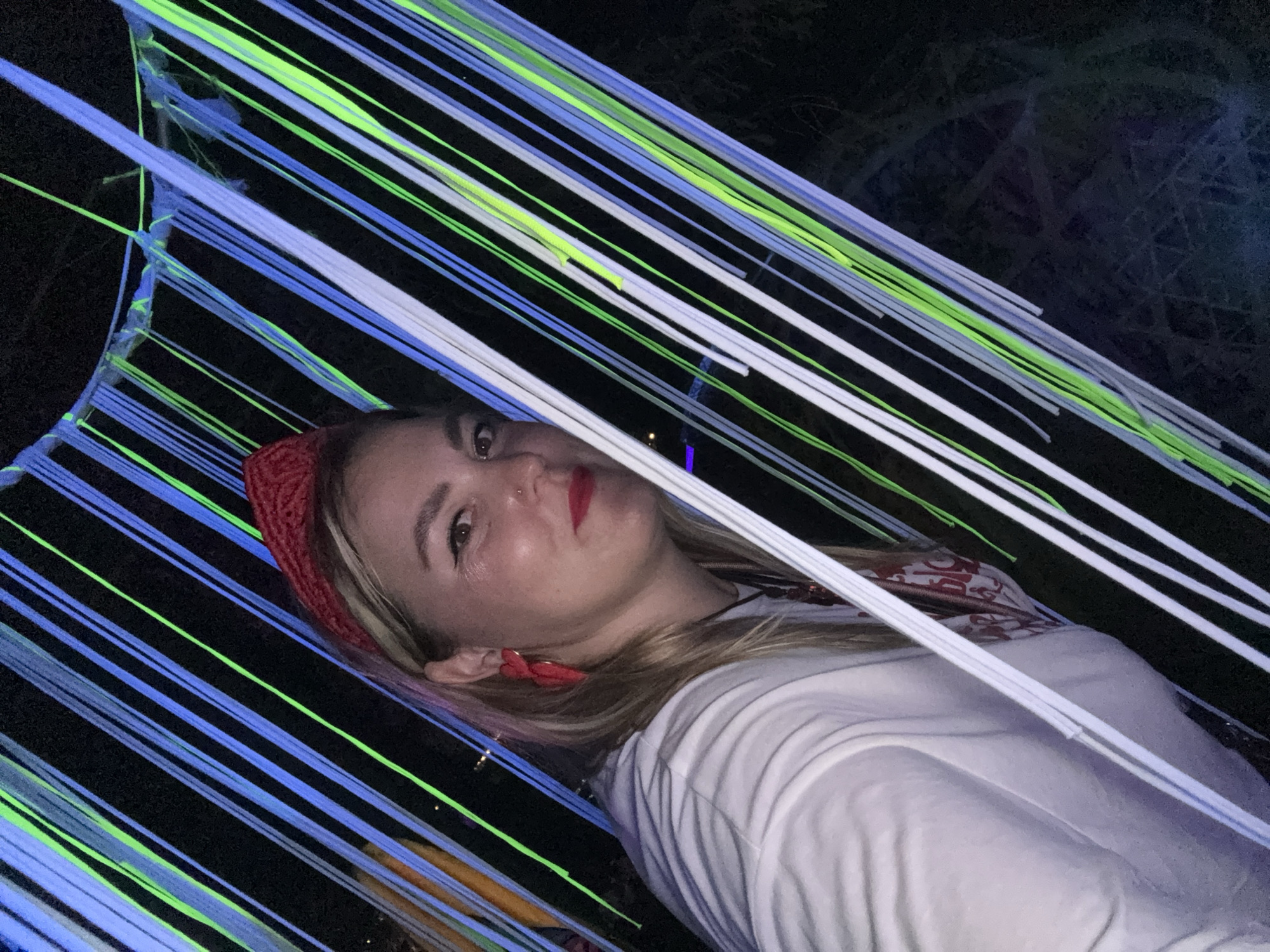 A woman with blonde hair, wearing a red bandana and a white shirt, is seen in a nighttime outdoor setting. She is adorned with large red earrings and red lipstick. She is looking towards the camera with a slight smile. The background is illuminated by strings of neon blue, green, and white lights, creating a vibrant and festive atmosphere. The exact location in Myl'niki, Russia, is not discernible from the image.