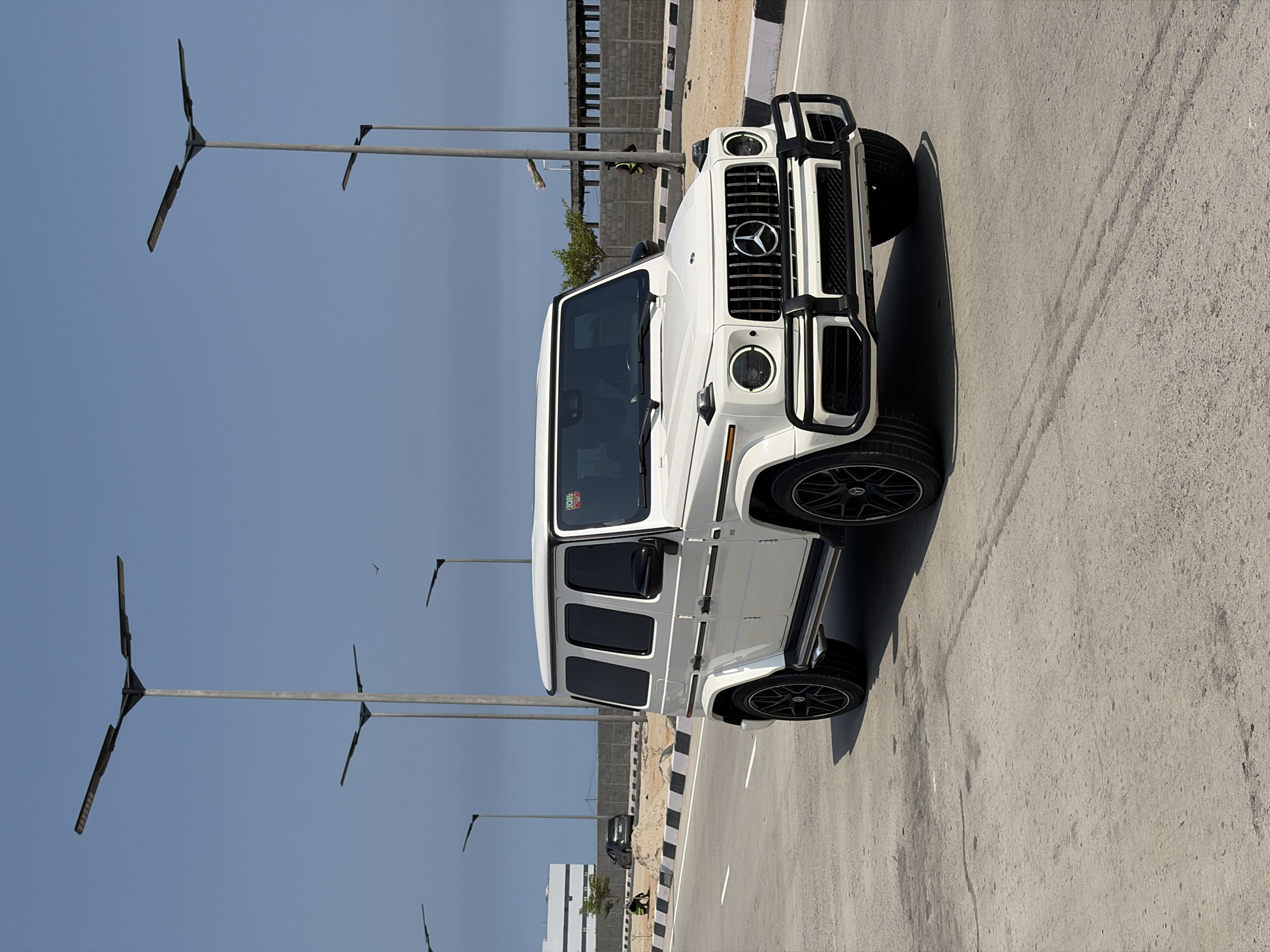 A white Mercedes-Benz G-Class SUV is parked on a gray asphalt road in Alaguntan, Nigeria. The vehicle is positioned near a curb with black and white markings. Several modern streetlights with horizontal bars extend along the road's edge. In the background, there's a partially constructed or unfinished building, possibly a bridge. A person is visible near the building. The sky is clear and blue. Other buildings and vehicles are further in the background.