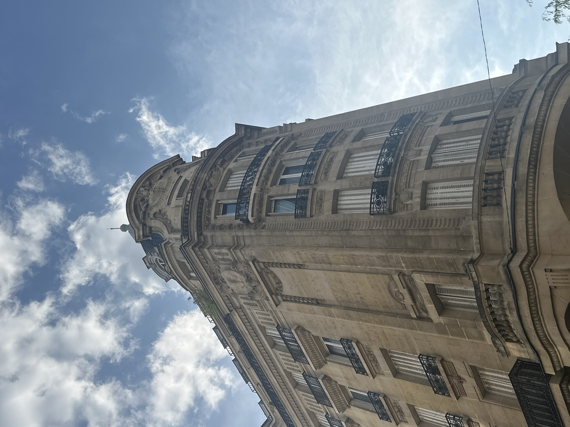 The image captures a close-up, low-angle view of the ornate facade of a Parisian Haussmann-era building, specifically the curved corner of an apartment building. The building, constructed from light-colored stone, features intricate architectural details, including decorative moldings, relief sculptures, and elaborate balconies with black wrought-iron railings. Multiple floors are visible, each with evenly spaced windows, many equipped with closed white shutters. The building's curved structure draws the eye upwards towards a cloudy blue sky. A thin, dark wire is visible diagonally across the sky on the right side of the frame. Vegetation, likely from rooftop gardens or trees, is subtly present on the upper levels of the building. The scene is devoid of any visible people or vehicles, emphasizing the architectural grandeur of the Parisian streetscape.