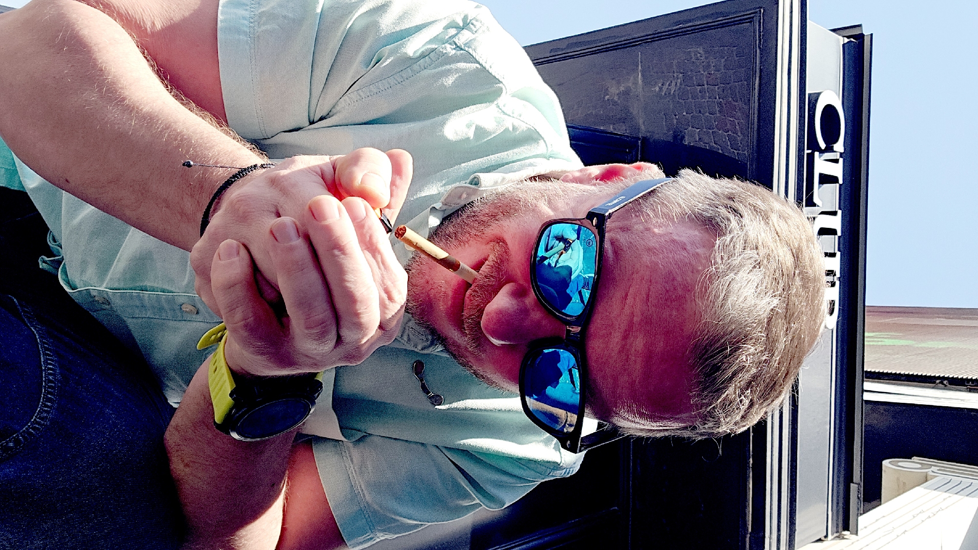 A man is smoking a cigarette outdoors on a sunny day. He is wearing blue jeans, a light blue button-down shirt, a black and yellow watch, and sunglasses with a blue reflection. The man is holding the cigarette between his fingers and close to his mouth. Behind him is a dark-colored door and some white signage. The location is Moscow, Russia.