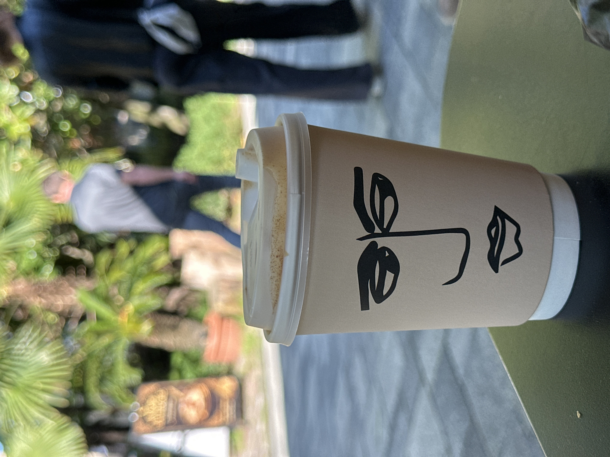 A coffee cup with a minimalist face design sits on a dark green surface, possibly a table. The cup is light brown with a white lid. The face design is black and stylized. The background shows blurred figures of people and lush greenery, including palm trees. The blurred elements suggest an outdoor setting. Given the context of Sochi, Russia, it is plausible that the photograph was taken in a cafe or public space within the city.