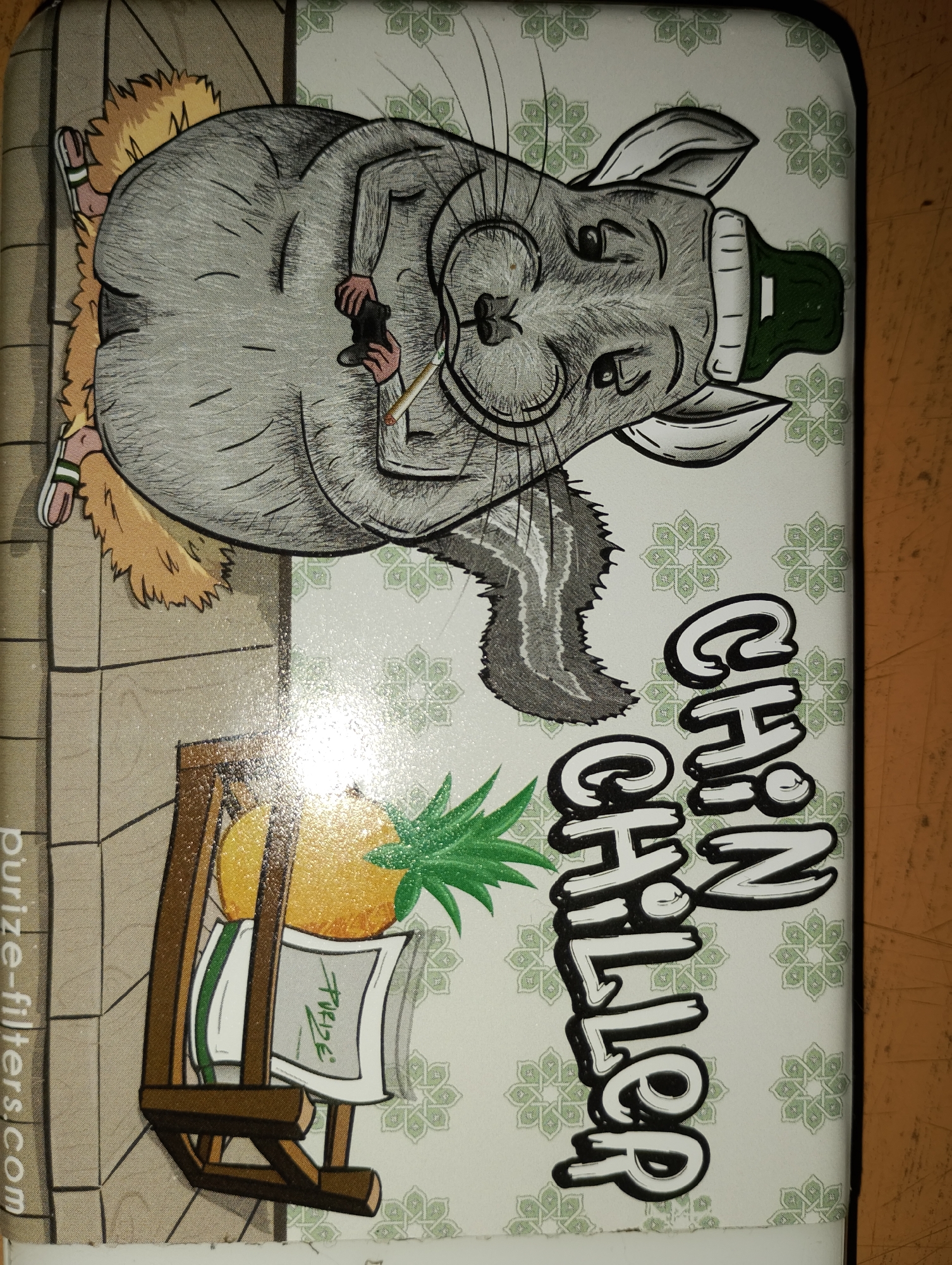 A digital illustration depicts a cartoon chinchilla lounging on its back, wearing a green beanie and holding a lit cigarette. The chinchilla appears to be on a fluffy, brown surface. To the left of the chinchilla, a wooden chair is placed in front of a white surface adorned with a repeating green floral pattern. A pineapple with green leaves sits atop the chair, resting on what appears to be a white cloth with green lettering that reads "Purize." The chinchilla is positioned on the upper right side of the image. Vertically oriented text in bold black and white letters spells out "CHILLER" on the right side of the image, overlapping the chinchilla and the floral pattern. A small section of a website address, "purize-filters.com," is visible on the lower left edge of the image. The background is a light-colored surface, possibly a table or countertop, with a textured appearance. The lighting suggests the image was taken in an indoor environment.