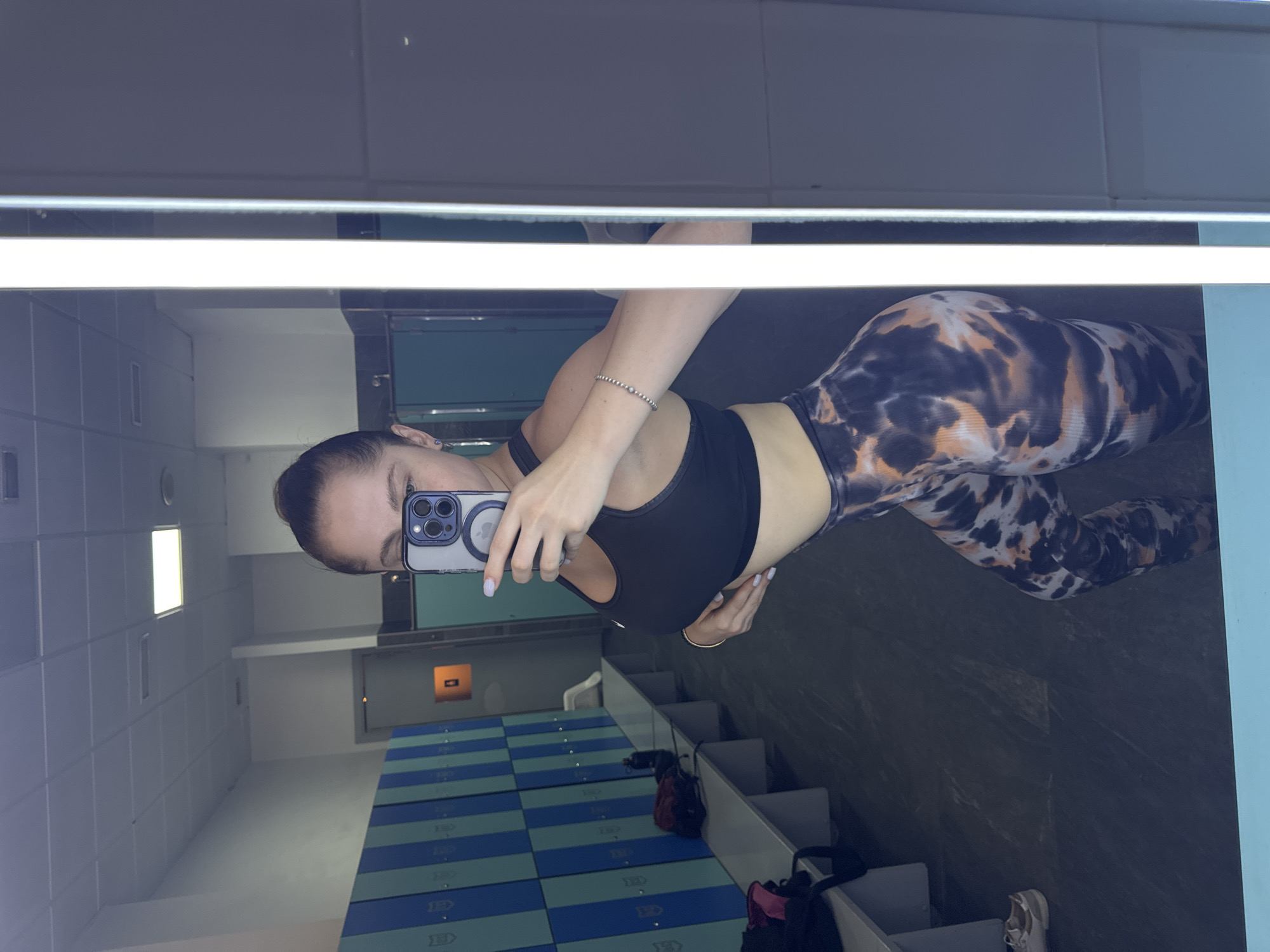 A young woman is taking a mirror selfie in what appears to be a locker room or gym. She is wearing a black sports bra and patterned leggings with a black, orange, and white tie-dye design. She is holding a smartphone in her right hand, taking the picture, and her left hand is on her hip. A silver bracelet adorns her left wrist. The background shows tiled walls, lockers, and a bright fluorescent light fixture overhead. In the reflection, a portion of a blue and green checkered floor is visible, along with some gym bags and shoes on shelves. The lighting suggests it could be daytime, and the setting is indoors. The woman's expression is neutral. No visible text is present in the image or its reflection.