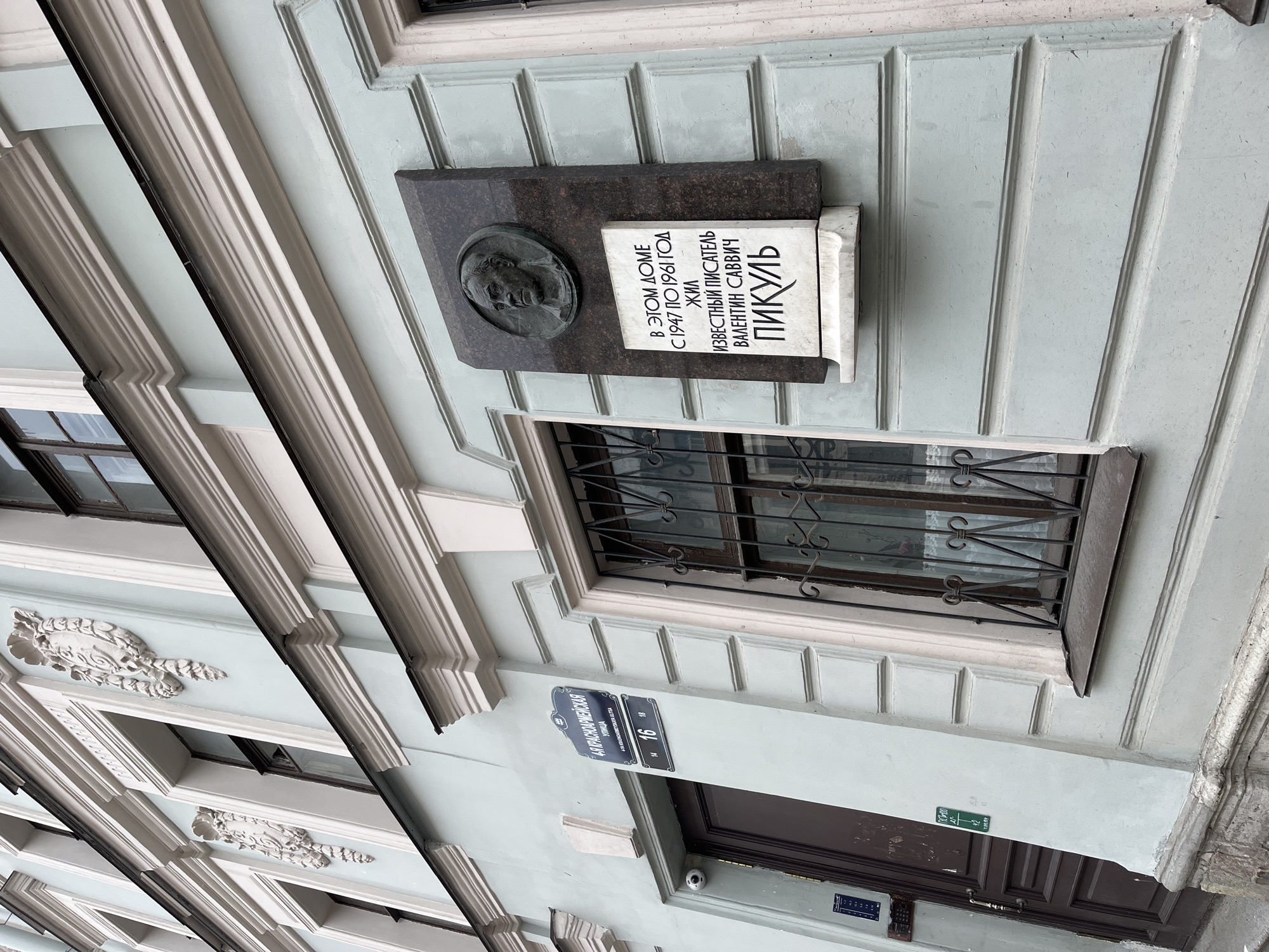 The image shows the facade of a building located at 16 Krasnoarmeyskaya Street in Saint Petersburg, Russia. On the wall, there is a memorial plaque dedicated to the writer Valentin Savvich Pikul, stating that he lived in this building from 1947 to 1961. A street sign is mounted below the window bearing the inscription "Krasnoarmeyskaya Street 16". The building is painted in a light blue color, and has a window with a decorative metal grille, with a dark-stained wooden door below the plaque. The facade features architectural details such as moldings. A surveillance camera is visible above the door.
