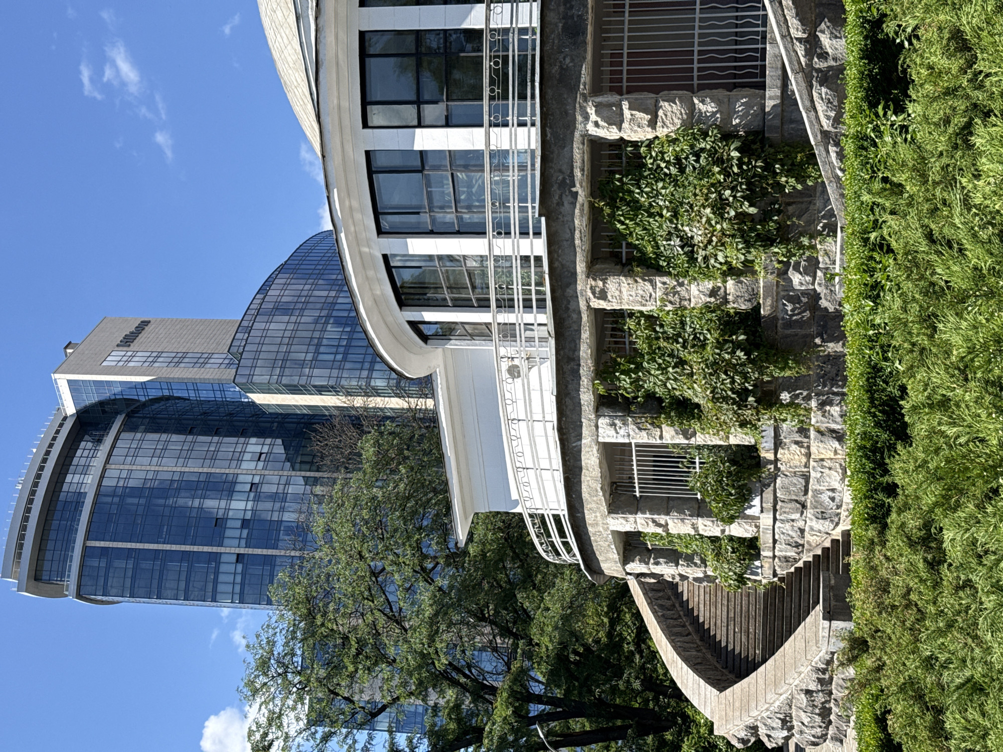 The image shows a curved stone structure with a staircase leading up to it, overgrown with ivy. Behind this structure, a modern glass-fronted building, the Hilton Kyiv, is visible, with a curved facade and a taller, rectangular section. The Hilton logo is clearly visible on the taller section. Trees with green foliage are present in the foreground and midground, obscuring parts of the buildings. The sky is blue with some scattered white clouds. The scene depicts an urban environment in Kyiv, Ukraine, with a blend of older architectural styles and modern skyscrapers.