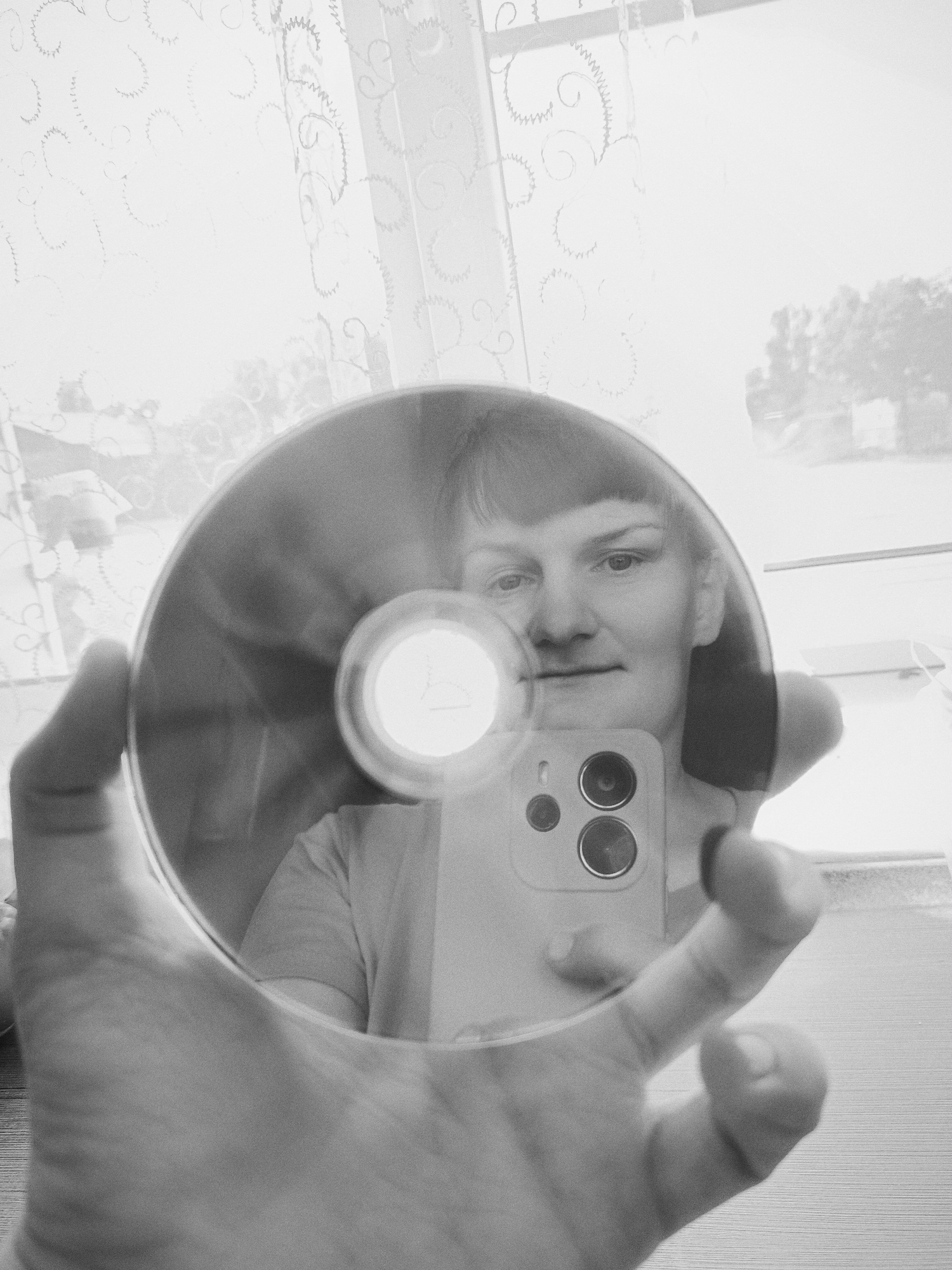A grayscale indoor photograph depicts a person holding a compact disc (CD) in their left hand, using it as a mirror to capture a selfie with a smartphone. The person's face is visible in the reflective surface of the CD, while the phone's camera lenses are also visible. The background consists of a window covered by a sheer curtain, beyond which trees are visible. The person's hand is in the foreground and appears large due to the close proximity of the camera. The location, Фоки, Russia is not discernible from the image.