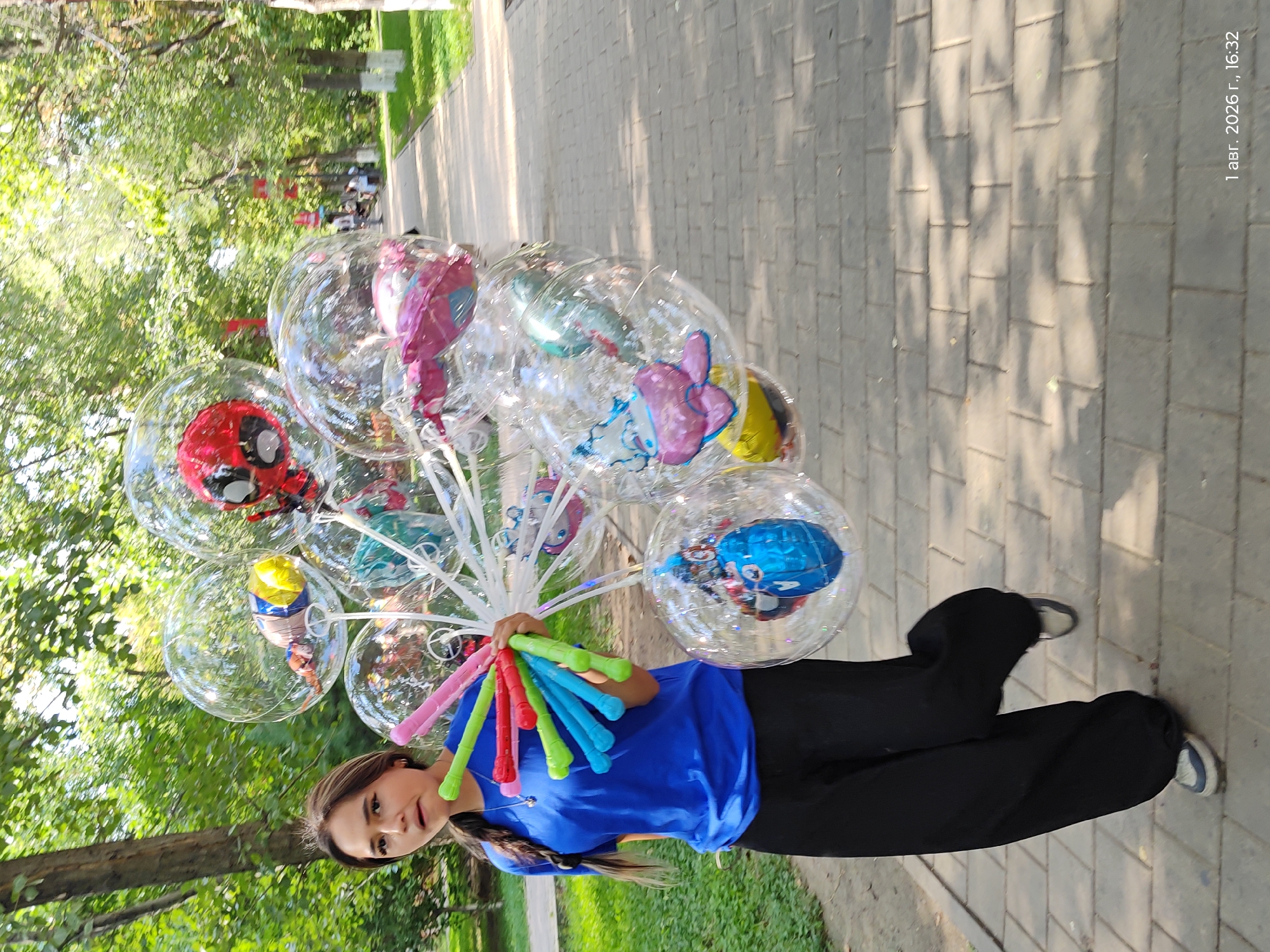 A young woman with long dark hair braided down her back, wearing a bright blue t-shirt and loose black pants, is walking along a paved walkway in a park. She is holding a large bunch of clear balloons, many of which contain smaller decorated balloons inside, including one shaped like Spider-Man and another like a pink jellyfish. The balloons are attached to colorful plastic sticks. The park setting features mature trees with green foliage, a grassy area to the left, and sunlight dappling the walkway, suggesting it is daytime. The walkway is made of light-colored rectangular pavers. In the background, other people can be seen walking in the distance, and there are red flags or banners visible. The timestamp in the lower right corner indicates "1 авг. 2026 г., 16:32," which translates to "August 1, 2026, 4:32 PM." This image was taken in Astana, Kazakhstan.