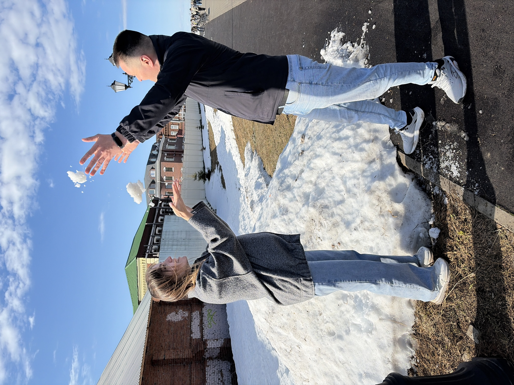The image shows a man and a woman in an outdoor setting with snow on the ground. The man, wearing a black jacket and jeans, is throwing a snowball. The woman, wearing a grey coat and jeans, is trying to catch it. They both wear sneakers. Behind the man, there is a building with a brick facade and lampposts. Behind the woman is a brick building with a corrugated metal addition and a blue sky with clouds above. The setting may be the city of Sarapul, Russia.