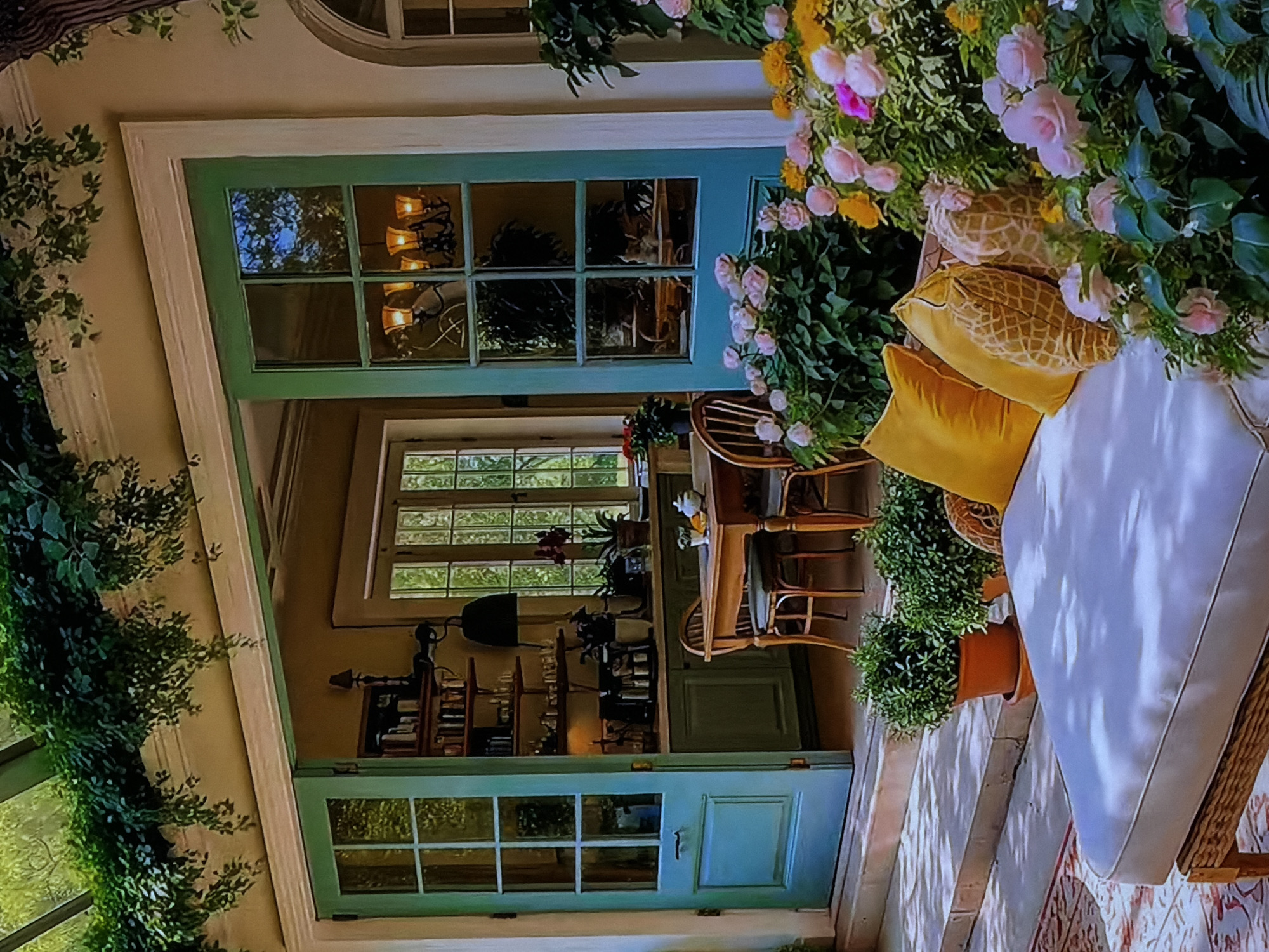 This image captures a vibrant and inviting indoor-outdoor living space, likely a patio or veranda adjoining a house in Sofiivska Borshchahivka, Ukraine. The scene is lush with natural elements and warm tones, suggesting a comfortable and relaxing atmosphere.
On the right side of the frame, a white outdoor sofa with thick cushions is adorned with several pillows in shades of yellow and a textured pattern, hinting at comfort. Overhanging the sofa is a profusion of flowering plants, primarily light pink roses and smaller yellow blossoms, creating a dense, verdant canopy. Below, potted green plants add to the natural aesthetic. The patio floor features patterned tiling or a rug, and parts of a stone or brick wall are visible.
The outdoor space opens into a well-lit interior through two sets of tall, teal-colored glass-paned doors. Through the upper set of doors, an interior room is visible, illuminated by a warm-toned chandelier with multiple shades. Reflections of foliage and sky are visible on the glass panes. Below, through the lower set of doors, another interior room appears, possibly a dining area, furnished with a wooden table and several chairs made of bamboo or rattan. Further back in this room, a bookshelf filled with books and decorative items is visible, along with a dark-colored lamp hanging above it. Multiple-paned windows are seen in the background of the interior spaces, allowing ample natural light. Green vines or ivy also climb the exterior wall of the house on the far left.
No people are visible, and no specific activity is taking place, but the setup suggests a peaceful area for relaxation or social gatherings. The combination of natural light and illuminated interior fixtures hints at either late afternoon or early evening. The overall impression is one of tranquility and warmth. There is no visible text in the image.