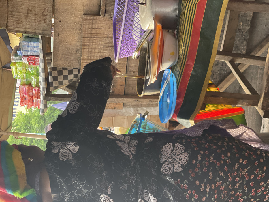 This image captures a vibrant and busy scene, likely within an outdoor market stall or informal shop, viewed from a slightly low angle. The image is rotated 90 degrees counter-clockwise; when viewed upright, it shows the following:
In the foreground, on the left side, a person is visible from the torso upwards, facing towards the right side of the frame. They are wearing a long-sleeved garment, possibly a dress or top, that is black with prominent white floral patterns. A colorful, rainbow-striped (red, orange, yellow, green, blue, purple) fuzzy hat or head covering is worn on their head, obscuring part of their face. Their left hand is raised, holding a thin, light-colored stick-like object, possibly for stirring, pointing, or handing something over. Part of another garment or the same person's layered clothing is visible on the far right, featuring a pattern of smaller, reddish-brown flowers on a dark background, with a checkered purple and white fabric at the neck/collar area.
The background reveals a rustic setup constructed from wooden planks and beams, forming shelves or a display structure. On the left side, various packaged goods are stacked, appearing to be small sachets or packets of products. Some white packets are clearly visible with the text "KLEENO", suggesting tissues, wipes, or a similar household item. Other packets are bright green and red. A black and white checkered cloth is draped over part of the wooden structure near these goods.
On the upper right side of the frame, a variety of kitchen or household items are displayed or stored. These include a purple plastic laundry basket, several metal pots or pans, and various plastic bowls and lids in colors such as white, orange, and blue. These items appear to be hanging or resting on wooden supports. Beneath these hanging items, a large piece of striped fabric with red, green, yellow, and black bands is draped over another wooden structure, possibly a mat or blanket.
In the mid-ground, behind the person and the stall structure, there is a glimpse of an outdoor environment with green foliage, indicating trees. A blue umbrella or canopy is also partially visible, along with a banner that has partial text visible, which appears to read "UNIVERSIT" on a blue background. The scene is brightly lit by natural daylight, suggesting it is daytime. The overall impression is one of an active, informal marketplace with various goods for sale.