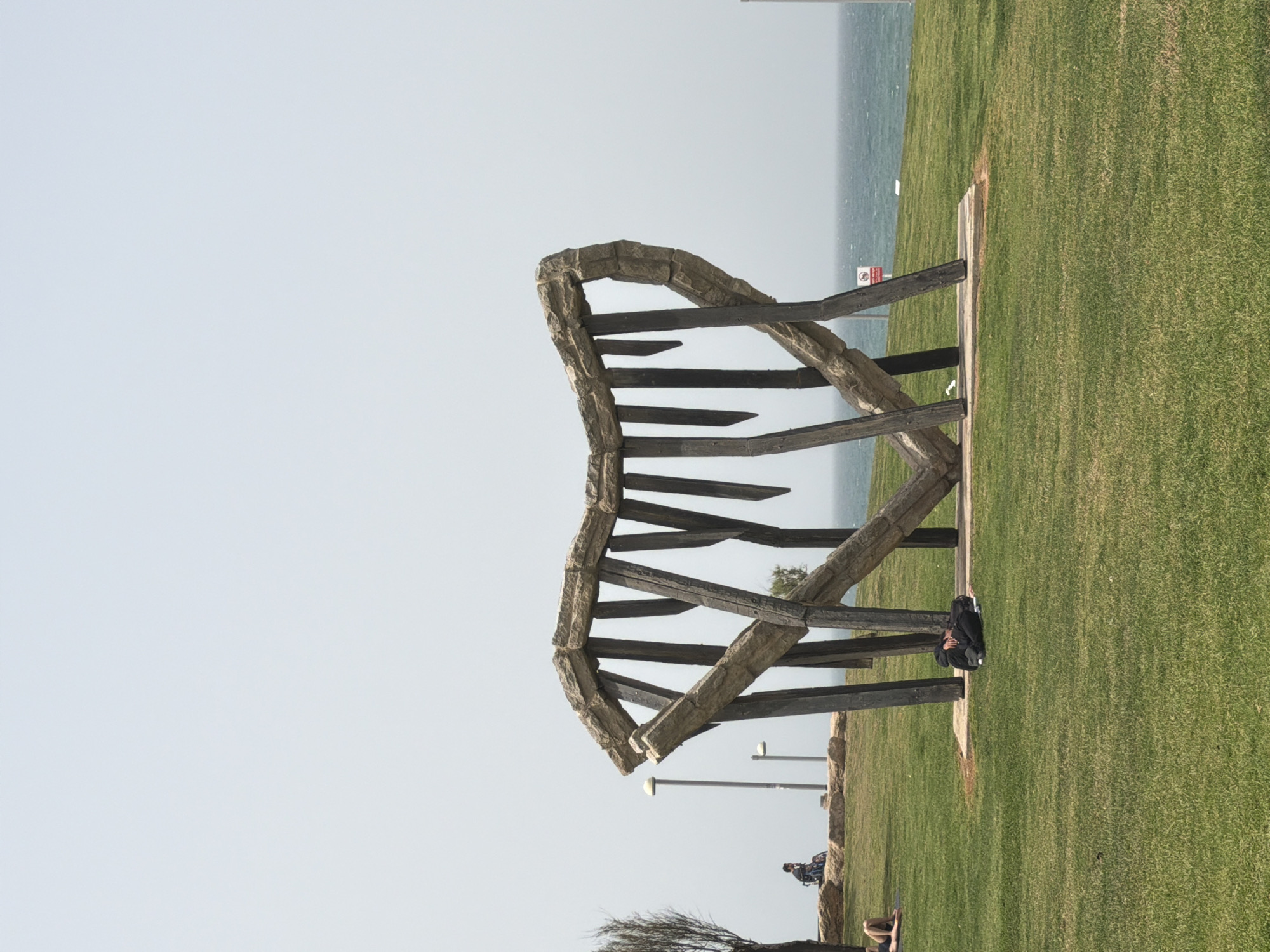 The image displays a large, abstract sculpture situated on a grassy area, overlooking a calm body of water under a clear, bright sky. The setting appears to be a coastal park or promenade in Tel-Aviv, Occupied Palestine.
The central subject is a prominent sculpture, composed of thick, rough-textured, light-colored stone or concrete forming an arched, rib-like framework, interlocked with dark, rectangular beams. This structure dominates the midground. Below the arch of the sculpture, a person is visible, resting or reclining on the grass. Further in the background, another individual can be seen on a bicycle or in a wheelchair near the water's edge.
The foreground and much of the midground consist of a wide expanse of well-maintained green grass. Along the edge where the grass meets the distant water, a paved pathway or border is discernible. Several modern streetlights are spaced along this path. The background features a vast, tranquil sea meeting a light blue sky. A small, partially obscured red circular sign with a diagonal line, typically indicating a prohibition, is visible near the water. The weather is clear and bright, suggesting daytime, and the overall atmosphere is peaceful and serene. A small, seemingly dormant tree or bush is located in the lower left foreground.