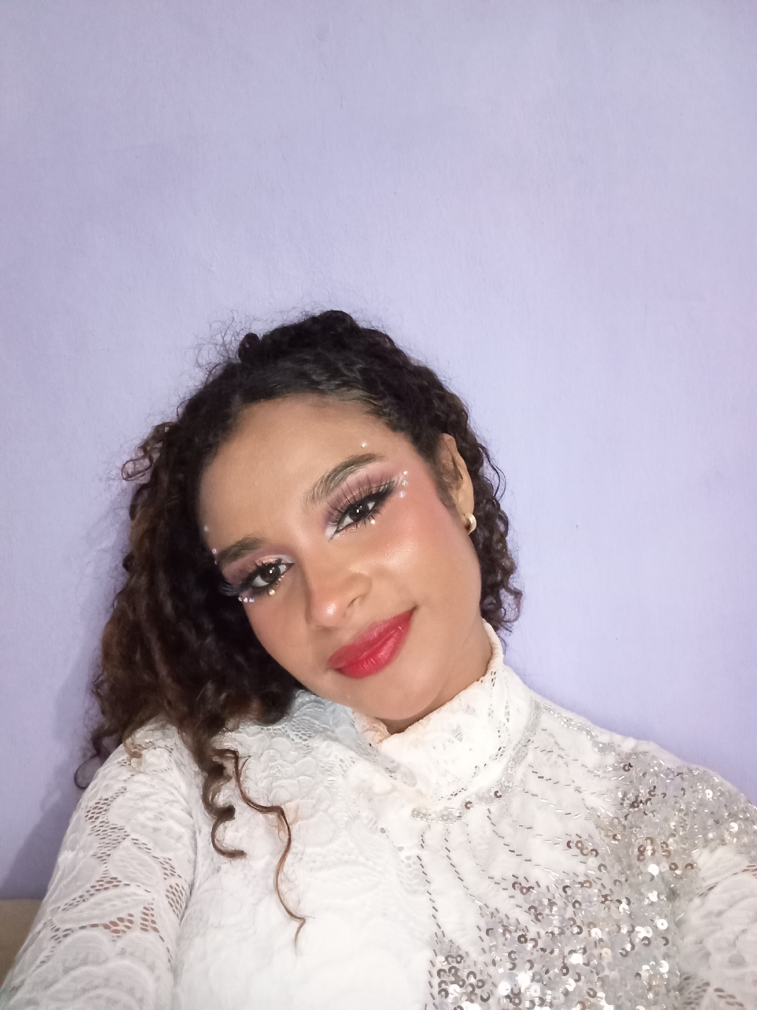 A woman with curly dark hair and light skin is in a selfie-style shot. Her makeup includes pink eyeshadow, prominent false eyelashes, and small, sparkling gems around her eyes and temples. She has red lipstick and appears to be smiling slightly. She is wearing a white, high-necked lace top with sequins on the right shoulder area. The background is a light lavender or purple color. There is no explicit indication of her location being in the city of Juan Griego, Venezuela.