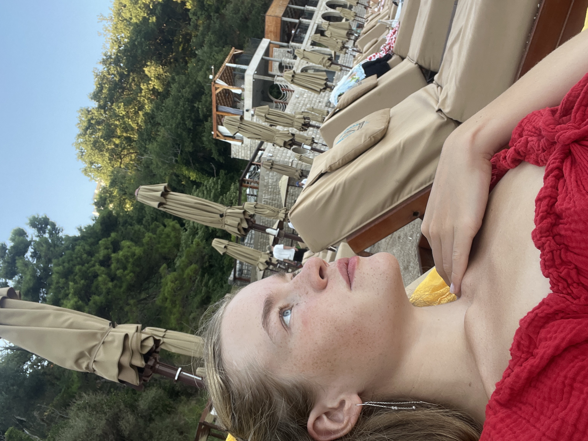 The image shows a young woman with blonde hair and freckles, wearing a red top, lying on a beach lounger. She is looking up and to her right, with her hand resting on her chest. In the background, there are rows of empty beach loungers and umbrellas, and a building with stone walls and a wooden roof. The scene is set in a coastal area with trees and a bright blue sky. This is likely a resort or a beach club in Budva, Montenegro.