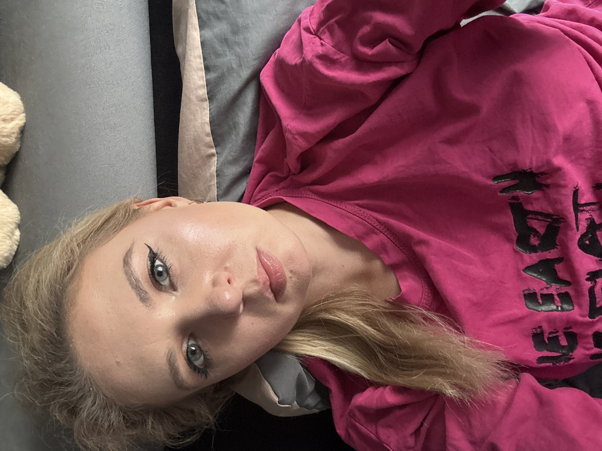 A woman with blonde hair and blue eyes is lying down, wearing a pink t-shirt with black text that reads "WE EART". The background appears to be a bed with grey and beige pillows. The lighting is soft and natural, suggesting an indoor setting. The woman is looking directly at the camera, with a neutral expression.