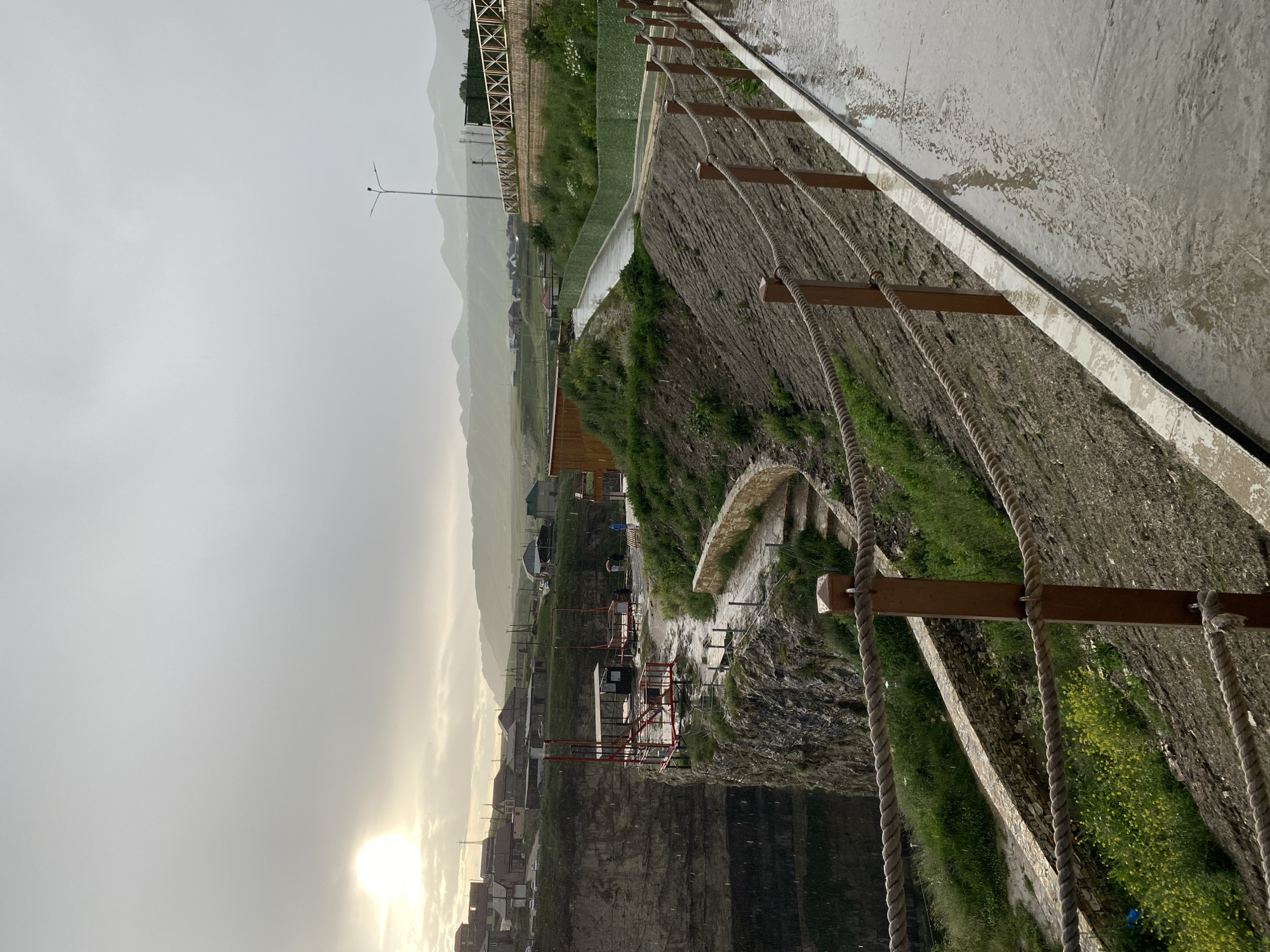 This image depicts the Khunzakh canyon, a natural attraction located in Khunzakh, Russia. The scene captures a cloudy day with a sliver of sunlight breaking through the overcast sky. In the foreground, a paved pathway, wet from rain, runs along a rope and wooden post barrier. The path is bordered by a steep, grassy slope.
Further into the image, a dramatic cliff face dominates the left side, with a structure built on its edge. This structure appears to be a platform with red railings and a covered area, suggesting it might be an observation point or part of an adventure activity. Several individuals, dressed casually, can be seen near this platform. Beyond the cliff and the platform, a village with houses and buildings is visible, nestled amongst rolling hills. The landscape is characterized by its rugged terrain and sparse vegetation, typical of a canyon environment. The overall atmosphere is somber due to the overcast weather, with a hint of subdued daylight.