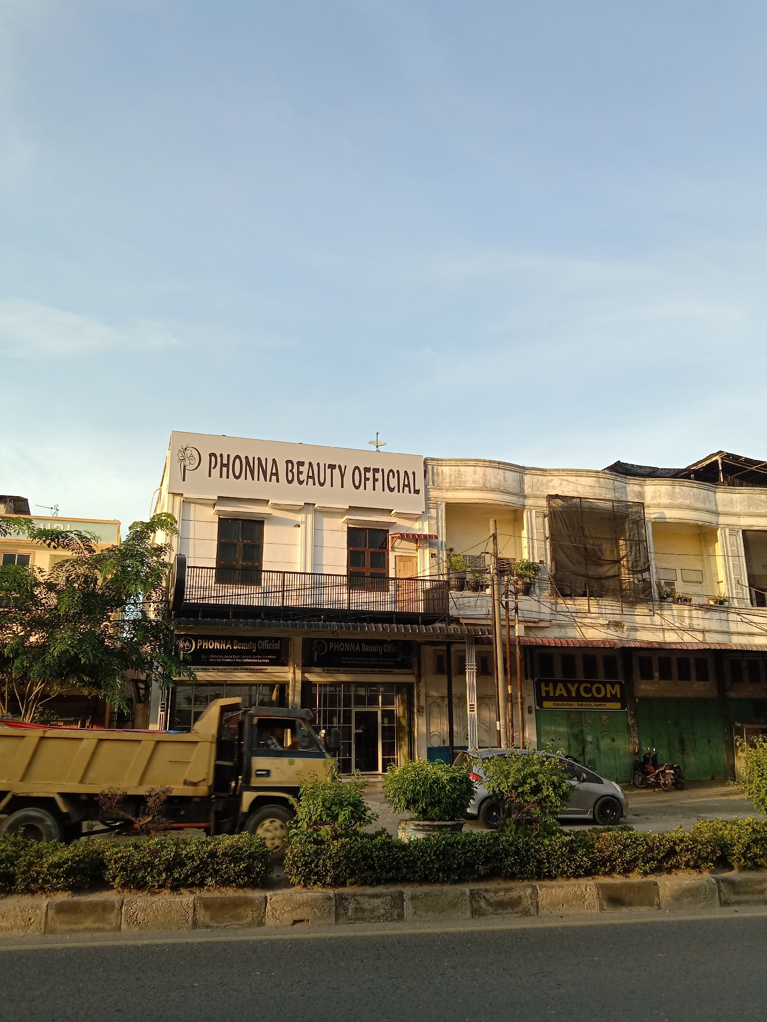 The image displays a street scene in Lhokseumawe, Indonesia, characterized by warm, directional sunlight suggesting either late afternoon or early morning. The sky is a clear, light blue with minimal cloud cover, indicating fair weather.
In the foreground, a road is visible, partially obscured by a median planted with green bushes and small trees. A yellow dump truck is captured in motion on the left side of the road, moving from left to right. Further along, a silver car is parked behind the foliage, and a motorcycle is discernible near a building on the right.
The background is dominated by a row of commercial buildings. The most prominent is a two-story white structure in the center, featuring a large sign on its upper facade that reads "PHONNA BEAUTY OFFICIAL." At street level, two smaller signs reiterate "PHONNA Beauty Official," advertising "Skincare, Body Care, Lipstik, lustre premium," and "One Set Premium dan Collection Lainnya." One sign also provides a detailed location: "Jalan Medan-Banda Aceh 5 mpang Buloh, Lhokseumawe."
To the right of the Phonnas Beauty building stands an older, beige two-story structure with decorative curved balconies, one of which is covered by a black mesh net. Adjacent to this, another building features green, garage-style doors and a yellow sign for "HAYCOM," specifying its products as "Kebutuhan Sekolah, Kantor" (School and Office Supplies). The overall scene portrays a typical commercial street with daily activity.