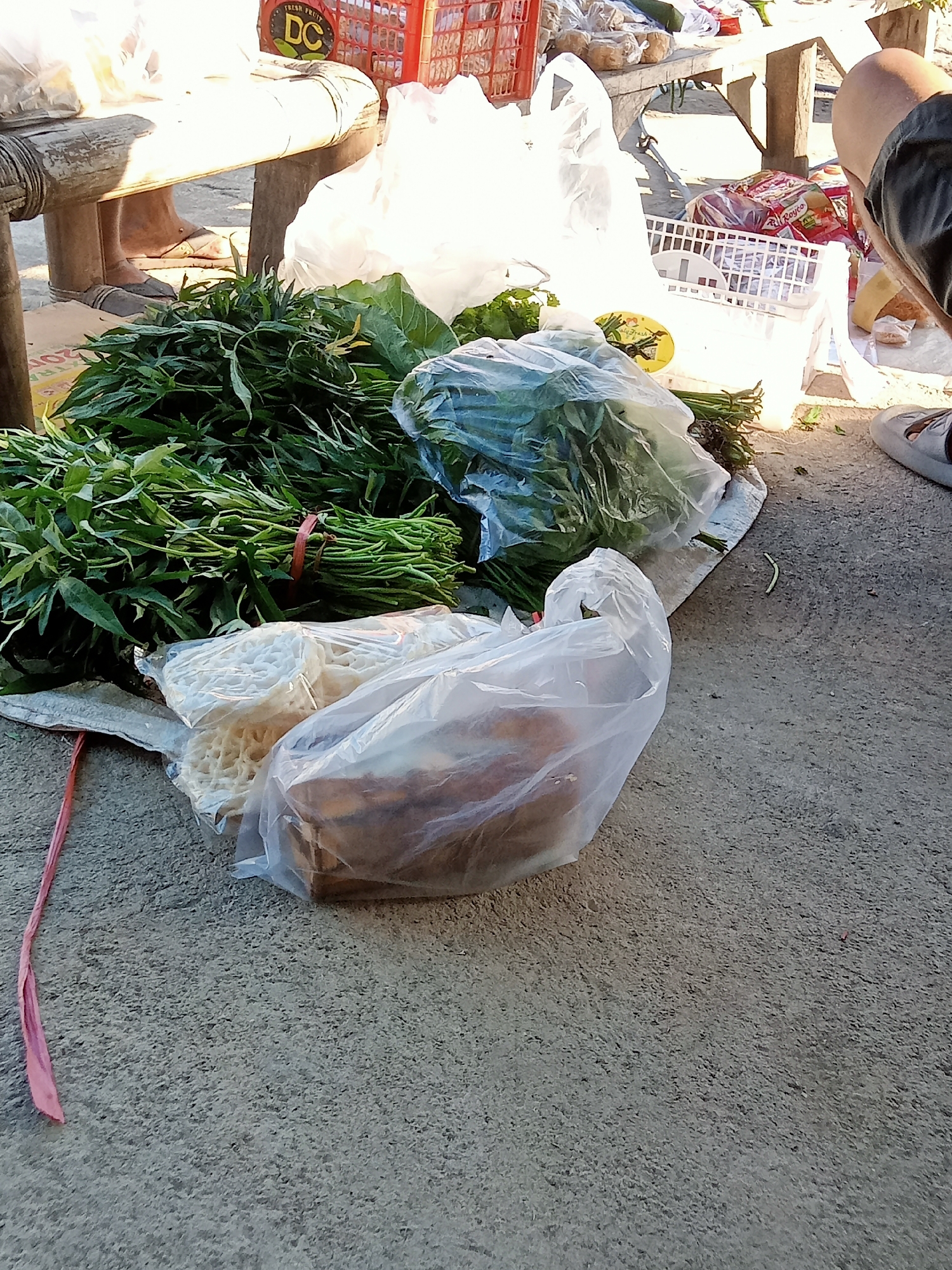 In Simo, Indonesia, a selection of fresh produce and bagged items is displayed on a concrete surface, likely at a market stall, alongside a wooden bench. A pile of green leafy vegetables, possibly water spinach, is tied with a red string and lies to the left. Next to it, a clear plastic bag contains white, circular crackers, and another bag holds what appears to be fried food items, possibly bananas or similar snacks. In the background, more produce, including what looks like cabbage, is visible in white plastic bags and a white basket. A person's legs and feet are visible, wearing casual footwear, near the wooden bench. Red plastic crates, one with "DC" branding, are also present in the background, indicating a commercial setting. The scene suggests a transaction or display of goods at a local market.
