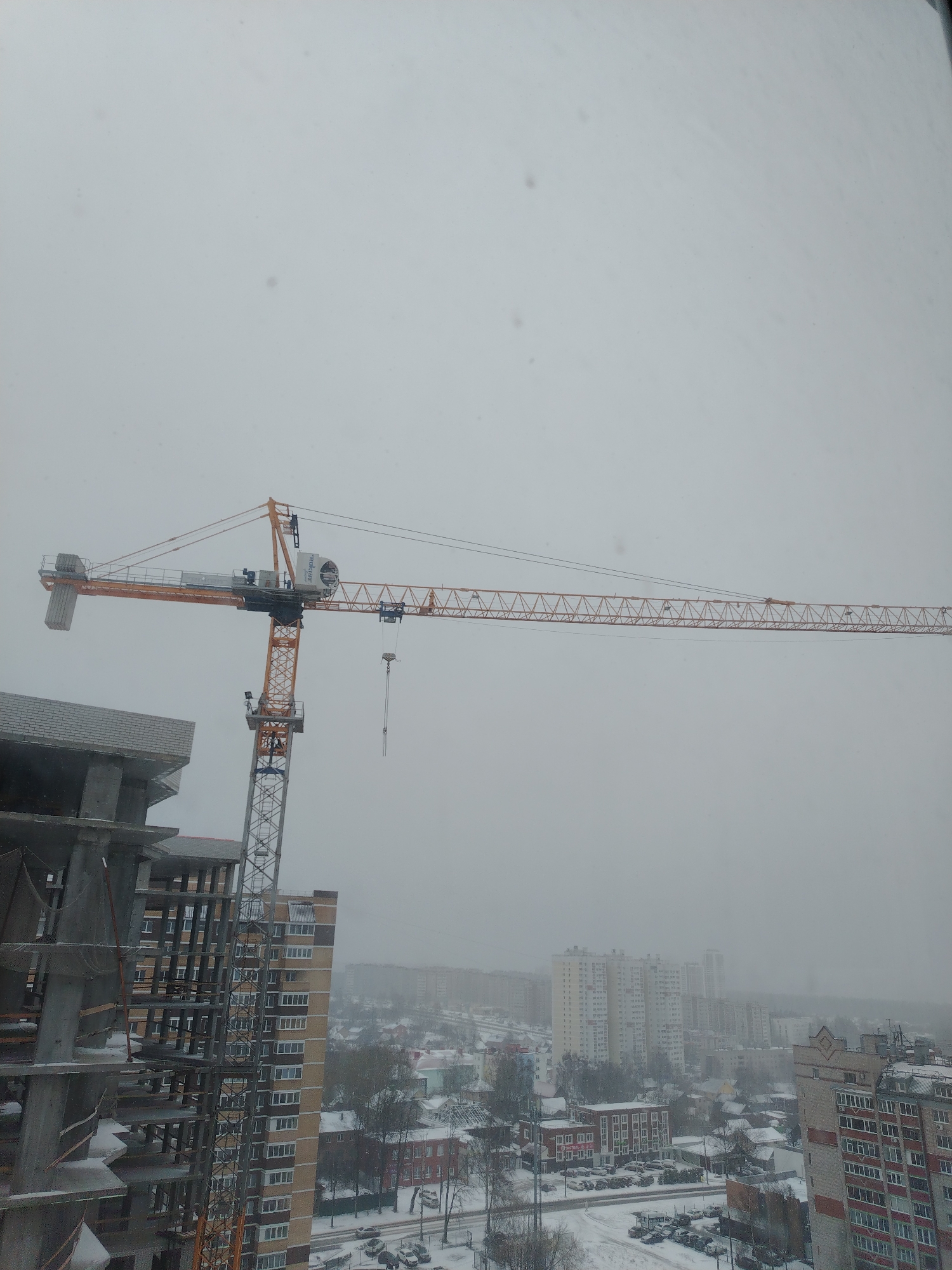The image depicts a cityscape in Izhevsk, Russia, under overcast, snowy conditions. A construction site dominates the foreground, featuring a high-rise building in progress alongside a large crane. The crane is positioned beside the building, its boom extending towards the right. In the background, a dense urban landscape unfolds with numerous multi-story residential buildings interspersed with patches of snow-covered ground. The atmosphere is hazy, obscuring distant details. No people are visible.