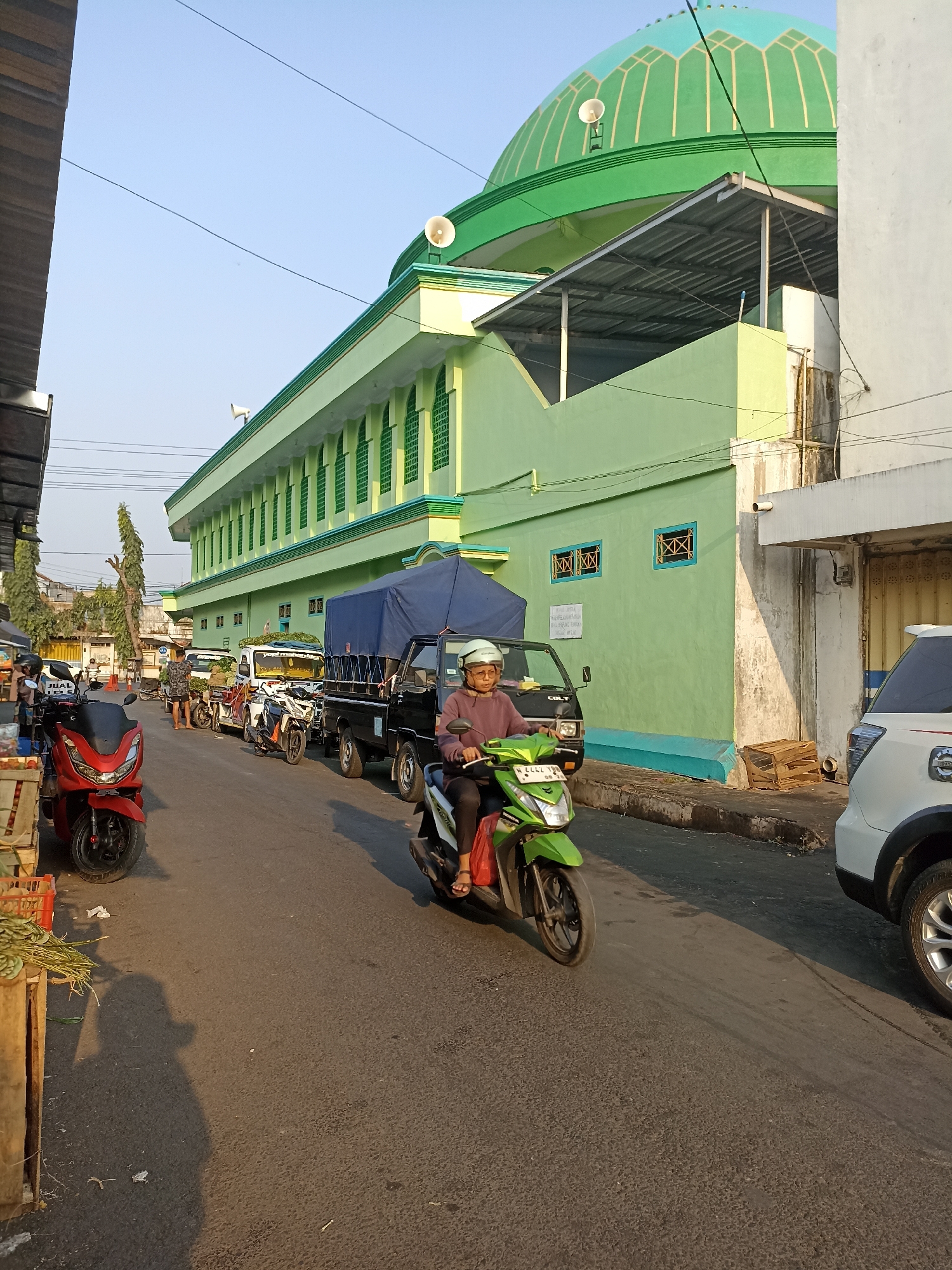 The image depicts a street scene in Tompokersari, Indonesia, on a clear, sunny day. A light green mosque with a large green dome dominates the background on the right side of the frame. The mosque features a long, light green building with evenly spaced, arched windows outlined in a darker green. A metal awning extends from the mosque building. On the left side of the street, a building with dark wood paneling is visible.
The street is lined with various vehicles. In the foreground, a person wearing a light helmet and a maroon shirt rides a green scooter. Behind the scooter, a red scooter is parked. Further down the street, several other scooters and small trucks are parked or moving. One black truck with a blue tarp covering its load is prominently positioned in the middle of the street. A white SUV is parked on the right side of the road, partially obscuring the mosque's structure.
On the left side of the street, a vendor's stall is visible with crates of produce, including what appears to be green vegetables or herbs. The road surface is asphalt, with some debris scattered on it. The overall impression is that of a busy street, likely a market area, adjacent to a religious building in an Indonesian city.