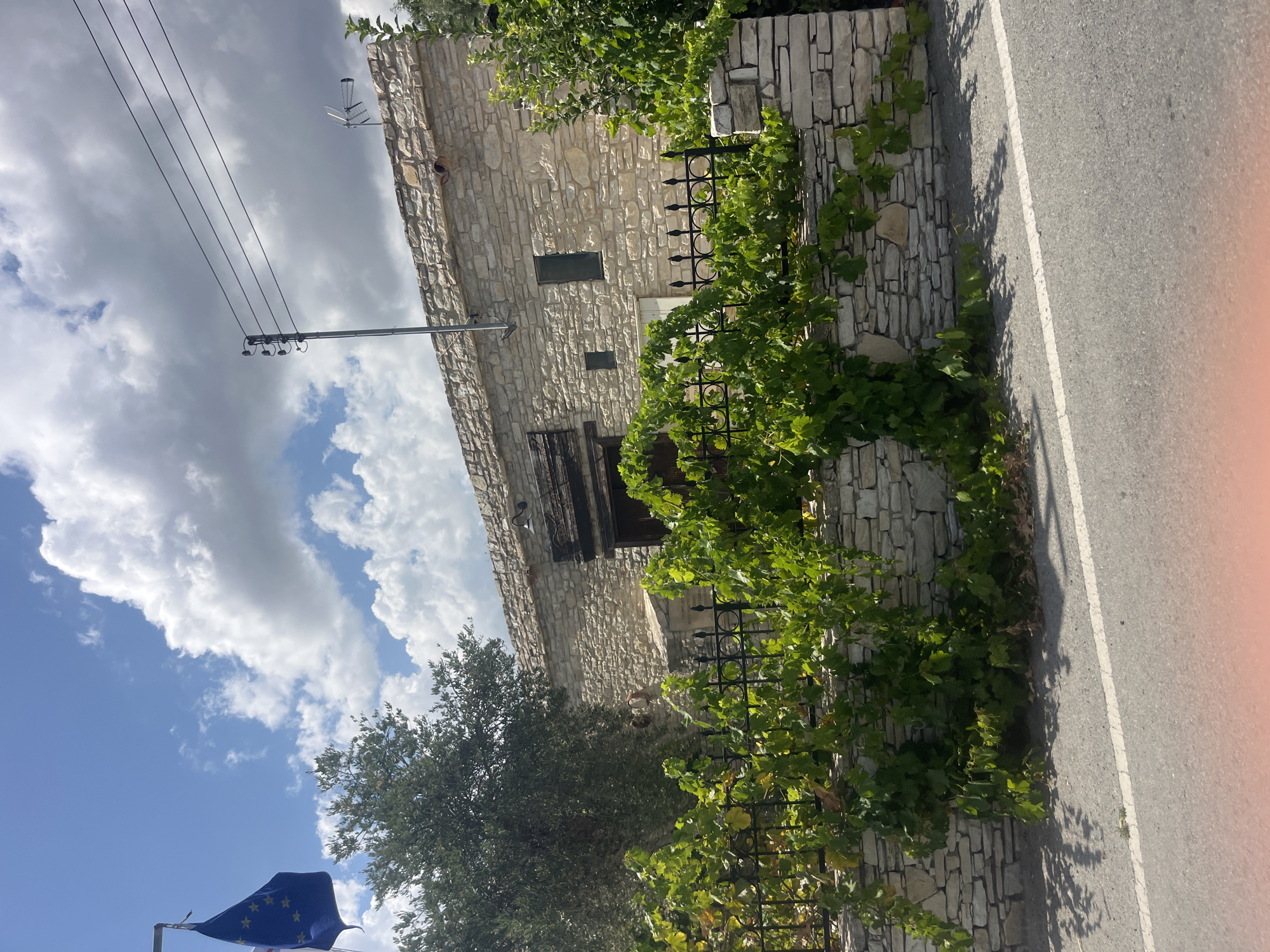 An external, medium-shot, eye-level, vertical, daytime photograph shows a stone building in Silikou, Cyprus. A European Union flag is visible in the top left corner, fluttering in front of a tree. The building, constructed with rough-hewn stones, has a single, small window and a larger wooden door. A black metal fence, topped with decorative spikes, runs along the front of the property, partially obscured by lush green vines. A paved road runs in front of the fence, and a white line marks the edge of the road. The sky above is a bright blue with fluffy white clouds. Power lines and a utility pole are visible above the building.