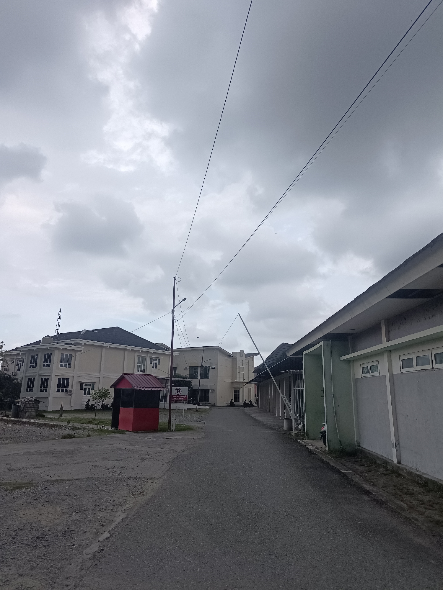 An overcast sky looms above a paved road winding through an institutional setting. On the left, a multi-story beige building with a dark roof is visible, featuring numerous windows. In front of it sits a small, red structure with a darker roof. To the right, a long, low building with a green section and a light gray wall stretches into the distance, also with multiple windows. Power lines crisscross the sky. A "No Parking" sign is posted near the road.