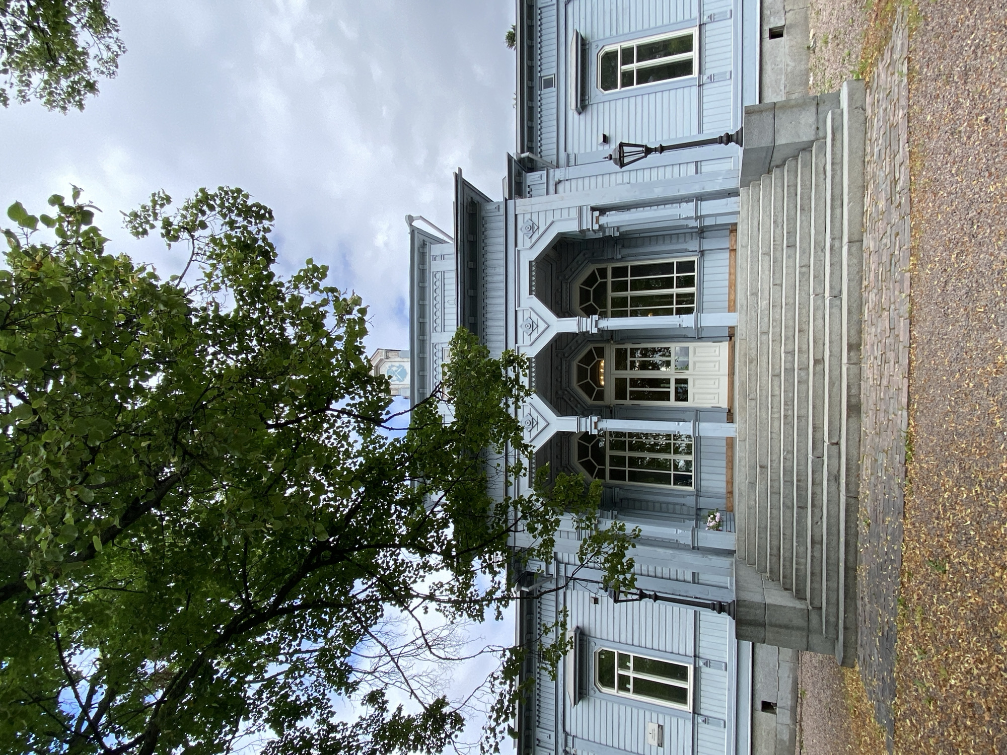 An exterior, eye-level, medium shot captures the facade of a light blue wooden building with a prominent central portico and a grand set of stone stairs leading up to the entrance. The building features white-trimmed windows and decorative architectural elements. Overhead, a large tree with green leaves partially obscures the sky, which is cloudy. The foreground consists of fallen leaves on a cobblestone path, with gravel visible beyond. The scene is set in Sortavala, Russia.