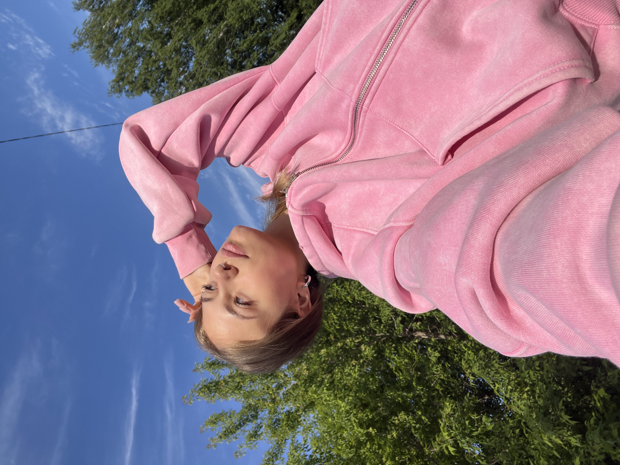 A young woman is the central subject of this rotated outdoor image, captured from a low-angle perspective, likely a selfie. She has light brown hair and light-colored eyes, with a nose piercing and ear piercings visible. She wears a relaxed, pink hooded sweatshirt (hoodie) made of a textured, possibly stonewashed fabric, featuring a silver zipper and front pockets. Her head is tilted, and one hand is gently touching her hair near her face, contributing to a calm, posed expression.
The background is dominated by a clear, bright blue sky with scattered thin, white clouds. Green foliage from trees is visible in parts of the background, particularly in the lower right and upper left corners of the rotated frame. A thin, dark line, possibly a power line, stretches horizontally across the sky in the middle-left section. The lighting indicates it is daytime. There is no visible text in the image. The setting is an outdoor area in Фоки, Russia.