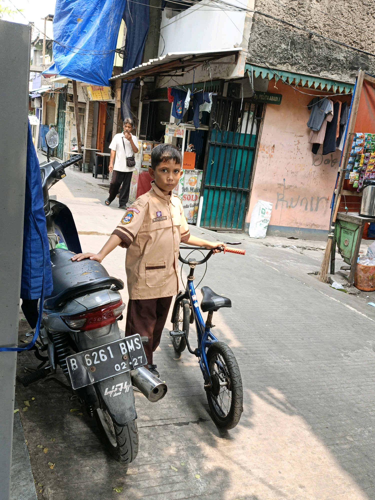A young boy in a tan school uniform stands in a narrow street, resting his left hand on a parked motorcycle and his right hand on the handlebars of a blue bicycle. The boy faces the camera with a neutral expression. The motorcycle is dark gray, with a license plate reading "B 6261 BMS 02-27." The bicycle is blue with red handlebar grips.
The street is paved with concrete, showing signs of wear. Buildings line both sides of the street, appearing to be shops or residences. Some buildings are adorned with colorful awnings or tarps. Clothes hang from lines on one building, and various goods are displayed on shelves outside another. A man in a white shirt and dark pants walks in the background, holding a phone to his ear. The scene is illuminated by bright sunlight, casting strong shadows on the ground. The environment suggests a densely populated urban area. The presence of the school uniform and the license plate format suggests this location is in West Jakarta, Indonesia.