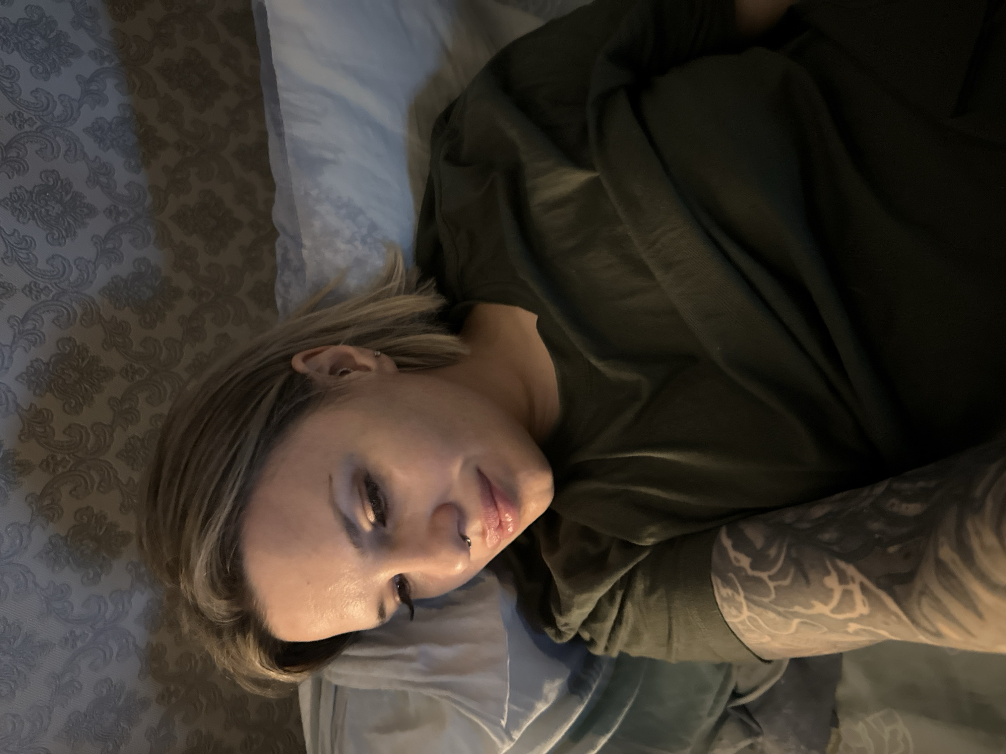 The image displays an adult female individual reclining indoors, possibly in a bedroom. There are no discernible visual cues within the image to specify a location such as Rostov-on-Don, Russia.
**Subject:** A female with light brown hair and blonde highlights, cut to approximately shoulder length, is positioned with her head resting on a light blue/grey pillow. Her eyes are directed downwards and to the left of the frame. She exhibits fair skin, a small silver hoop piercing in her nasal septum, and a small silver stud earring in her visible right earlobe. Minimal makeup is present on her eyes and lips.
**Attire:** She is wearing a dark olive green short-sleeved t-shirt. Her right arm, visible on the left side of the frame, is extensively covered by a full-sleeve tattoo featuring complex dark and light abstract or organic patterns.
**Environment:** The background consists of a wall covered in patterned wallpaper, characterized by light grey or beige damask-like motifs on a darker grey base. The bedding is of a similar light blue/grey tone to the pillow. The lighting is subdued, illuminating the subject's face primarily from the left, suggesting ambient or directional light from an unseen source.
**Actions/Interactions:** The individual is in a static, reclined posture, possibly observing an unseen object below her line of sight. No interactions with other persons or objects are evident.