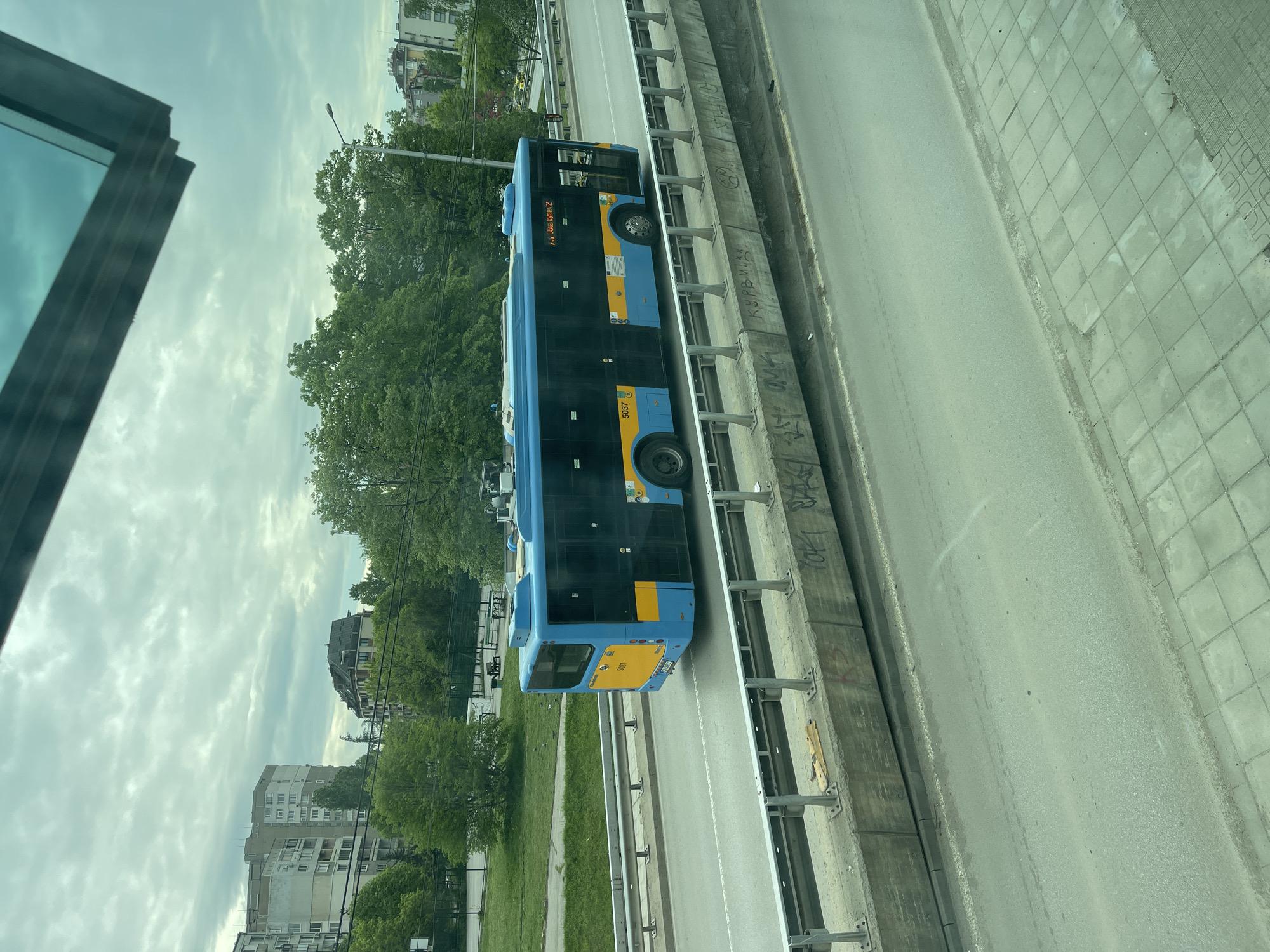 The image depicts a blue and yellow bus traveling on a road in Sofia, Bulgaria. The bus has the number 5037 printed on it. Trees and buildings are visible in the background. The photo was taken from a slightly elevated position, possibly from another vehicle or building. A pedestrian walkway is seen next to the road. Graffiti markings are visible on the concrete barrier beside the road.