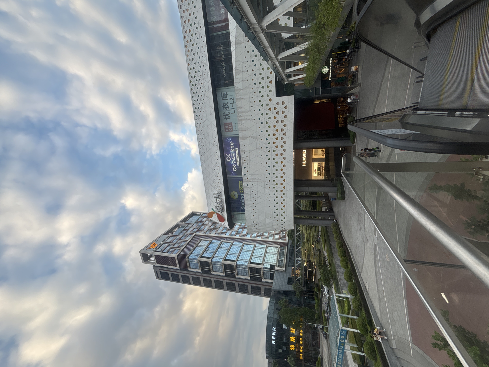 This is a medium-shot, eye-level photograph taken outdoors in the Liwan District, China, featuring a modern commercial complex under a cloudy sky. The complex includes a tall, multi-story building with a distinctive angular top featuring an orange logo, and a lower, wider structure with a white, patterned facade. Several prominent businesses are visible on the lower building, including "CK PAR KTV", "UNIQLO", "优衣库", "URBAN REVIVO", and "HUAWEI". A Starbucks Coffee shop is also identifiable.
In the foreground, an escalator descends, with a silver handrail in the lower left and right corners. Beyond the escalator, a paved plaza area is visible with several individuals walking and sitting. Two women are seated on a low wall to the left of the plaza. A covered structure with an "ENTER" sign is located further down the plaza. The scene is characterized by clean lines and modern architecture, suggesting a contemporary urban environment. The lighting indicates it may be late afternoon or early evening, with diffused light from the overcast sky.