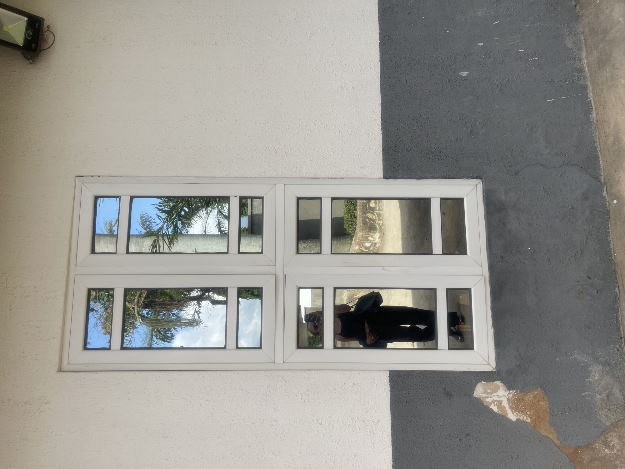 A person is reflected in a window on the exterior wall of a building. The building's wall is painted in two horizontal sections: a lighter color on top and a darker grey on the bottom. The window is a vertical multi-paned casement window with white framing. The upper panes reflect palm trees and a blue sky with clouds. The lower left pane shows a reflection of a person wearing black pants, a black tank top, and sunglasses, holding a dark bag and what appears to be a phone. Their feet, wearing dark sandals, are visible in the very bottom pane. The building appears to be located in Bukuru, Nigeria, as indicated by the context provided. The surrounding environment includes a rough concrete surface at the base of the wall, with some chipped paint and discoloration on the grey lower section of the wall. A security light is mounted on the upper right section of the wall, above the window.